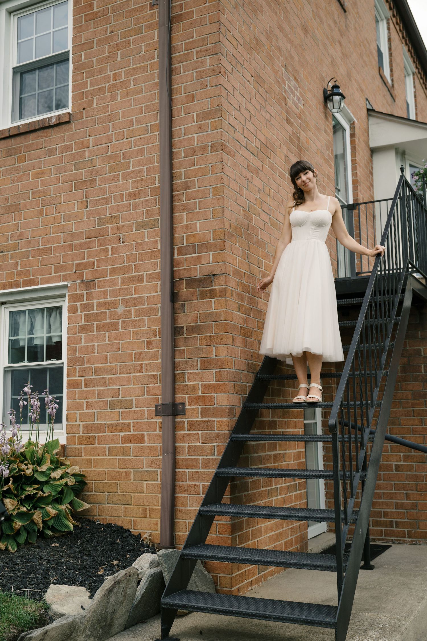 A bride in a tea-length white dress poses on exterior metal stairs of a brick building.