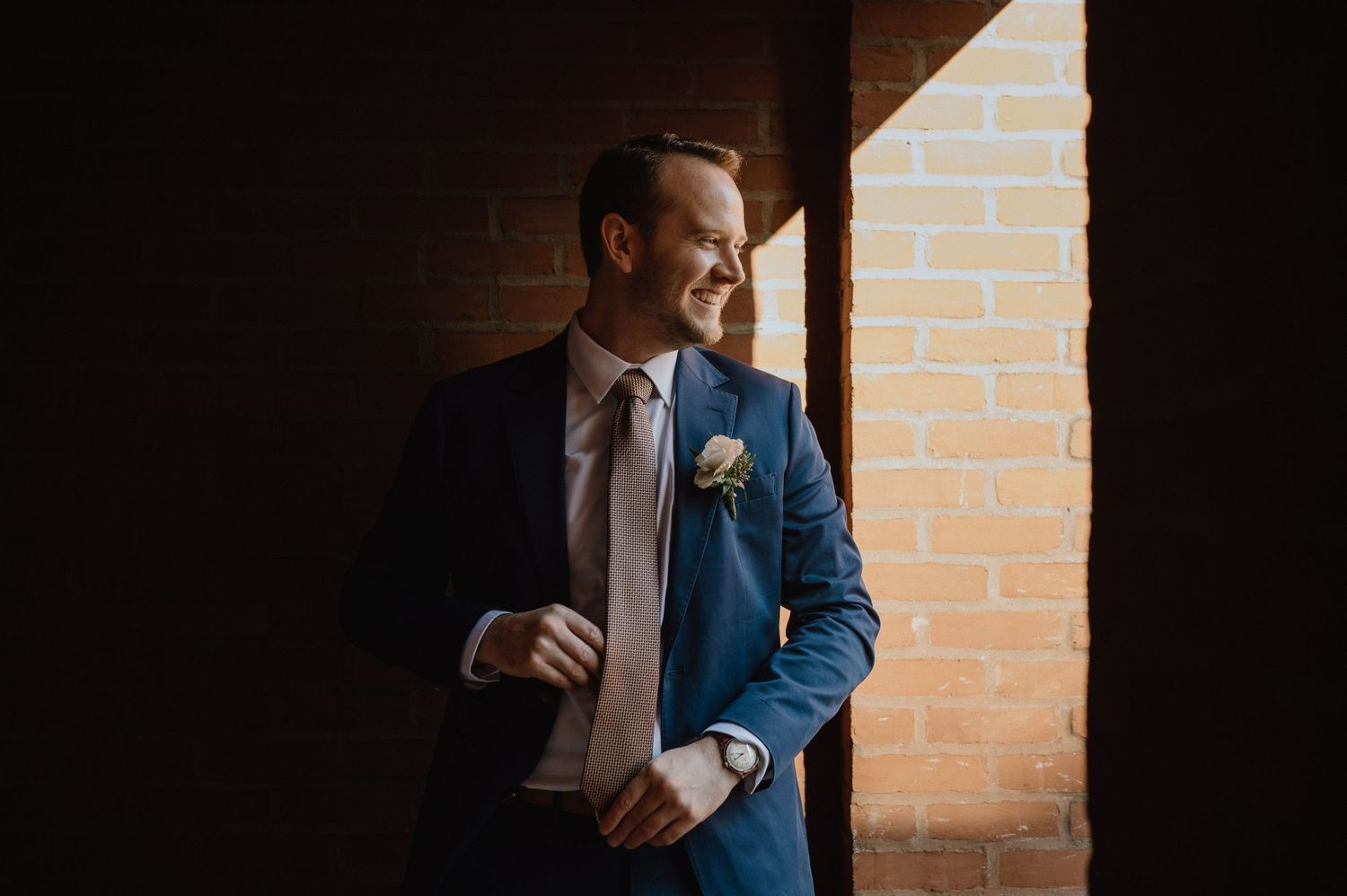 A person in a blue suit and boutonniere stands in dramatic window light against brick wall.