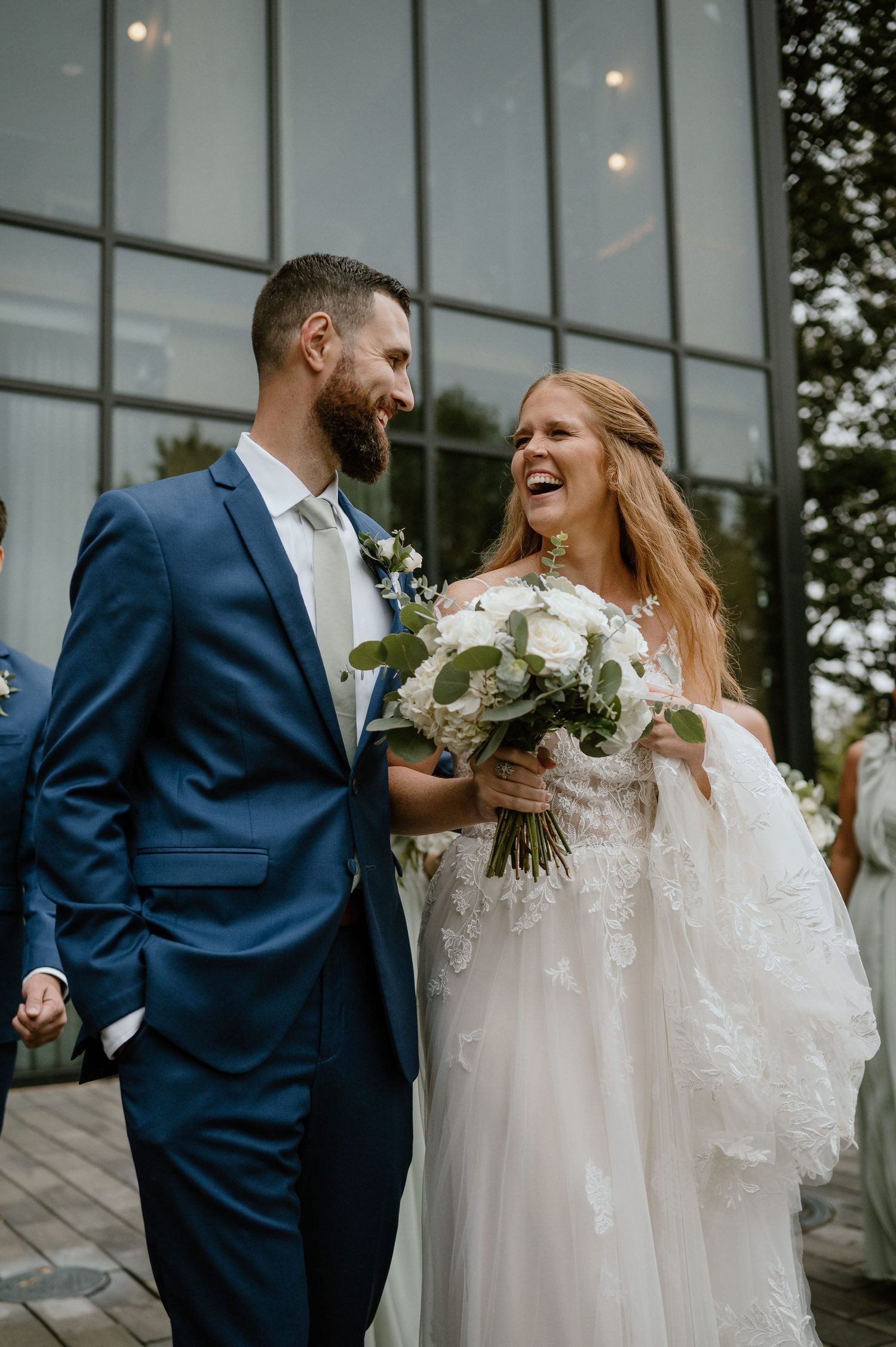 Candid moment of newlyweds laughing together outside modern venue with glass facade.