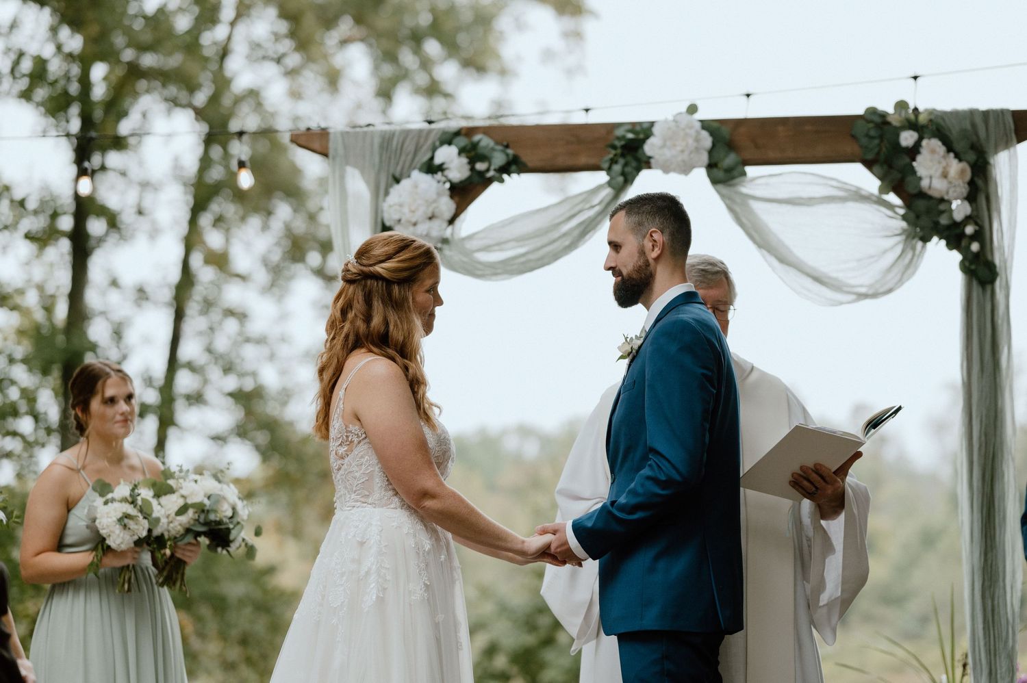 Wedding ceremony under wooden arch decorated with white fabric and floral arrangements.