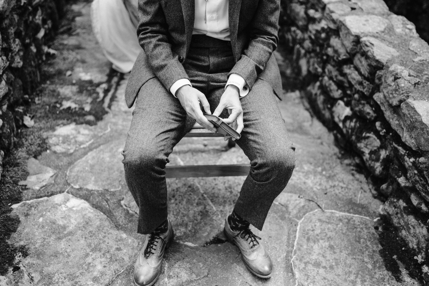 Close-up black and white shot of person sitting on stone wall holding phone.