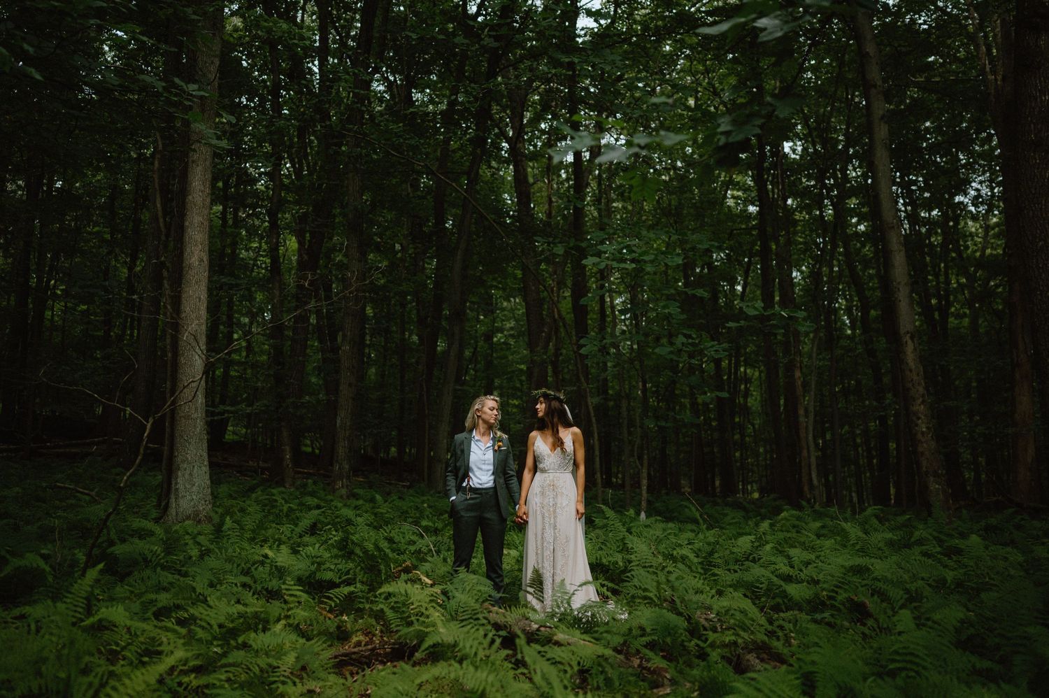 A couple stands together in a lush forest setting surrounded by tall trees and ferns.