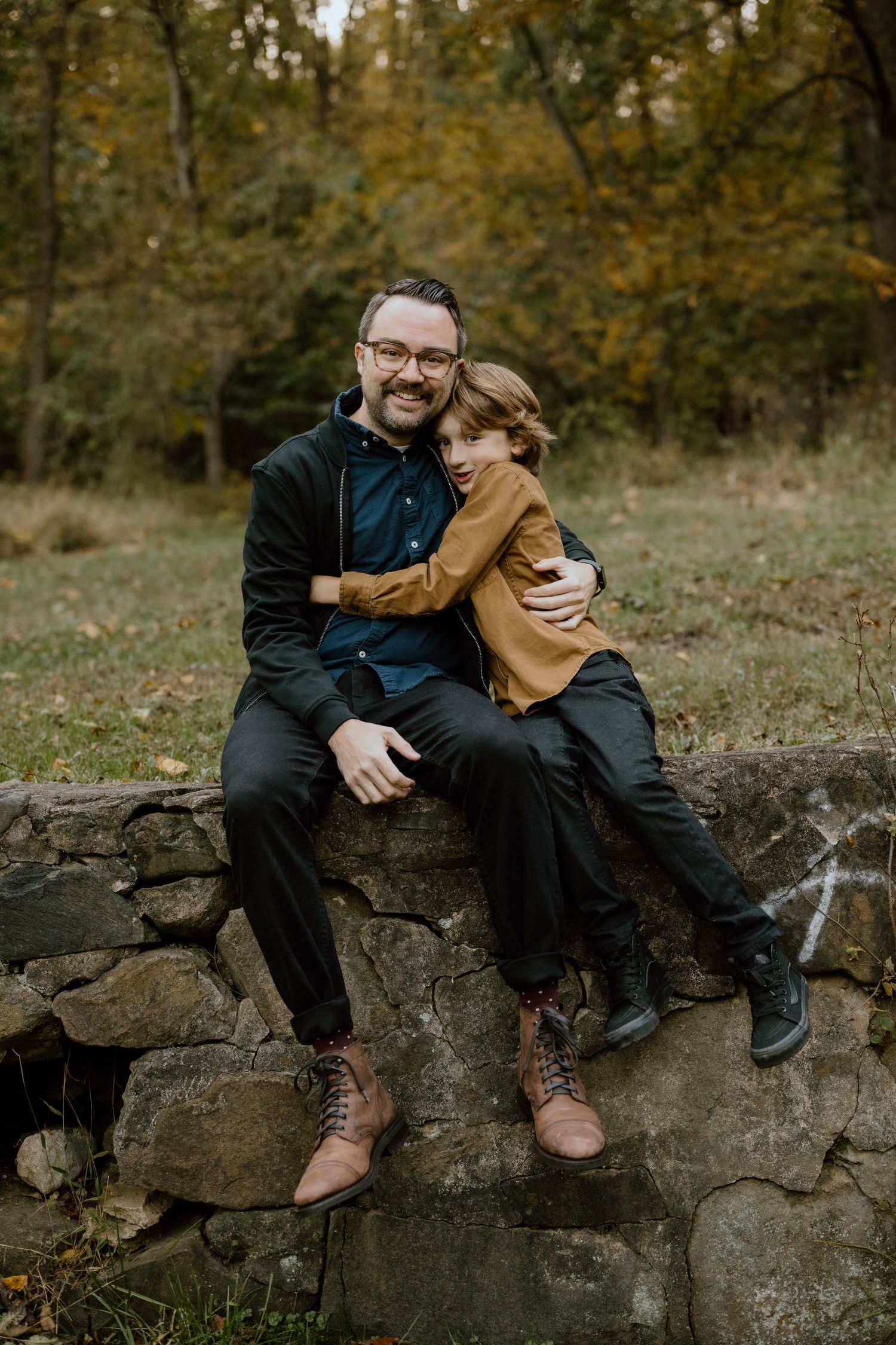 Two people share a heartfelt moment while sitting on a stone wall in an autumn setting.
