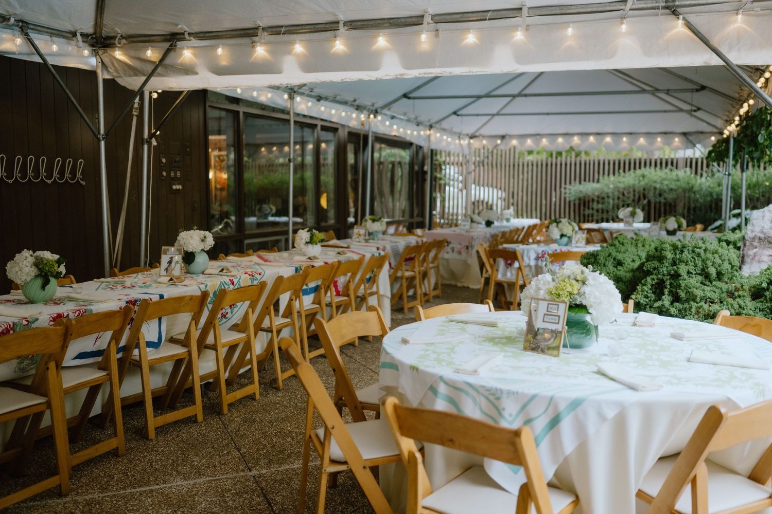Outdoor wedding reception setup with string lights and wooden chairs under white tent.