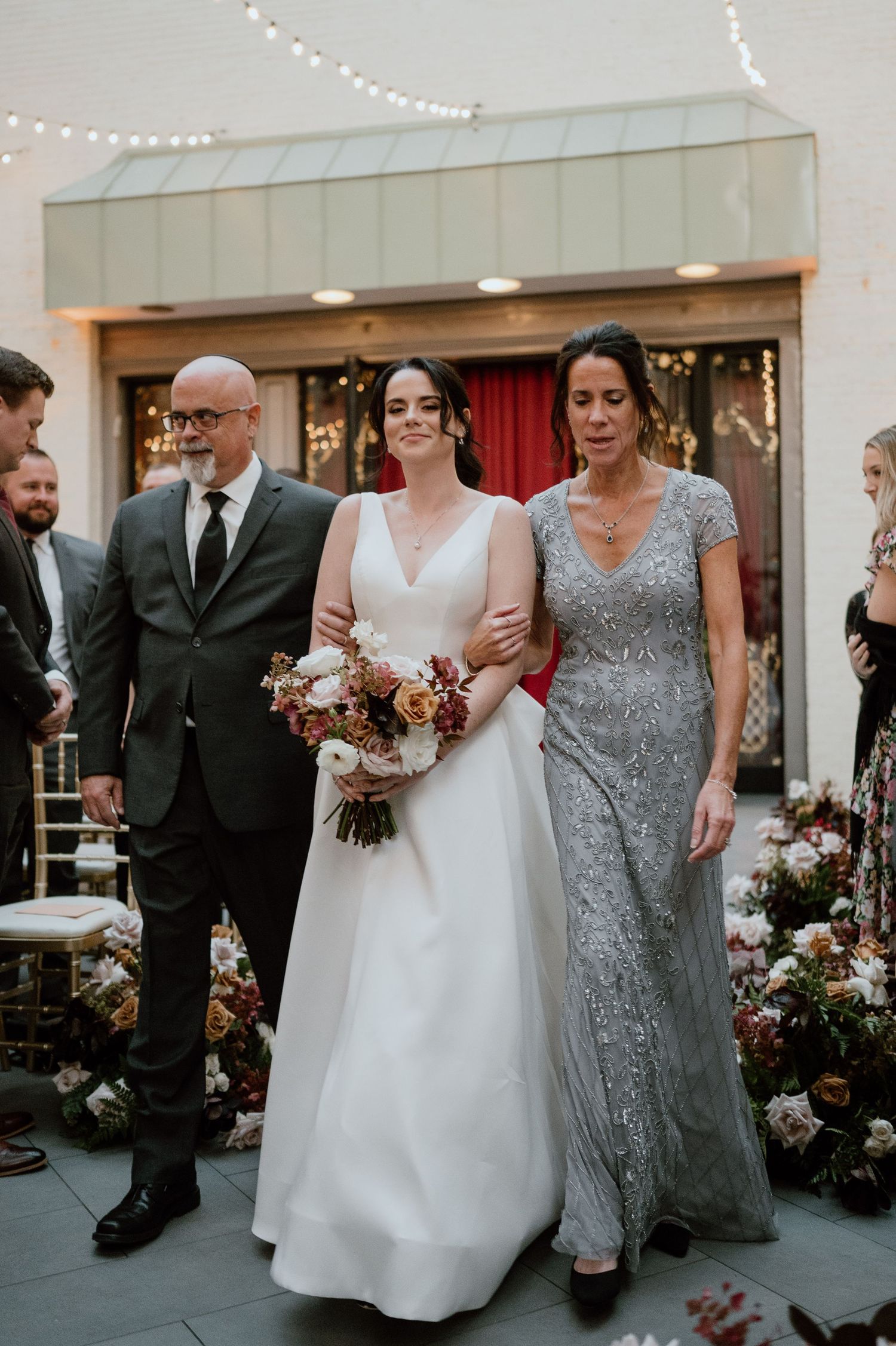 Wedding ceremony entrance with white dress and formal attire against floral decorations.