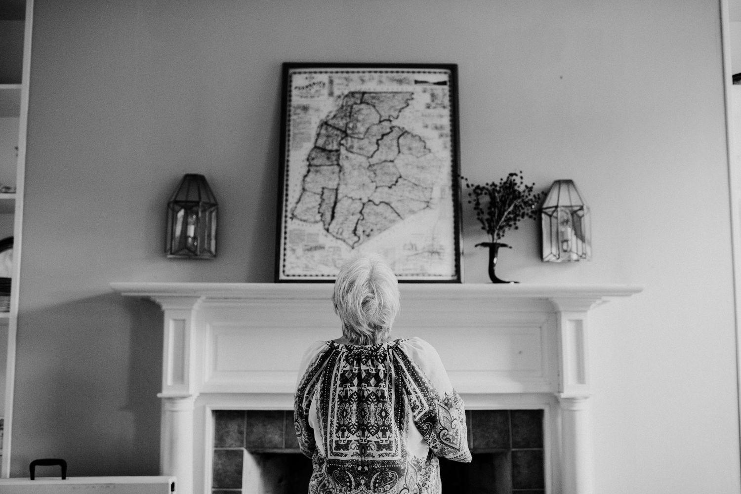 A black and white photo showing someone looking at framed artwork above a fireplace mantel.