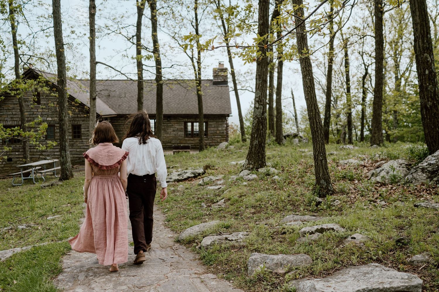 A couple walks toward a rustic stone cottage through a wooded path.