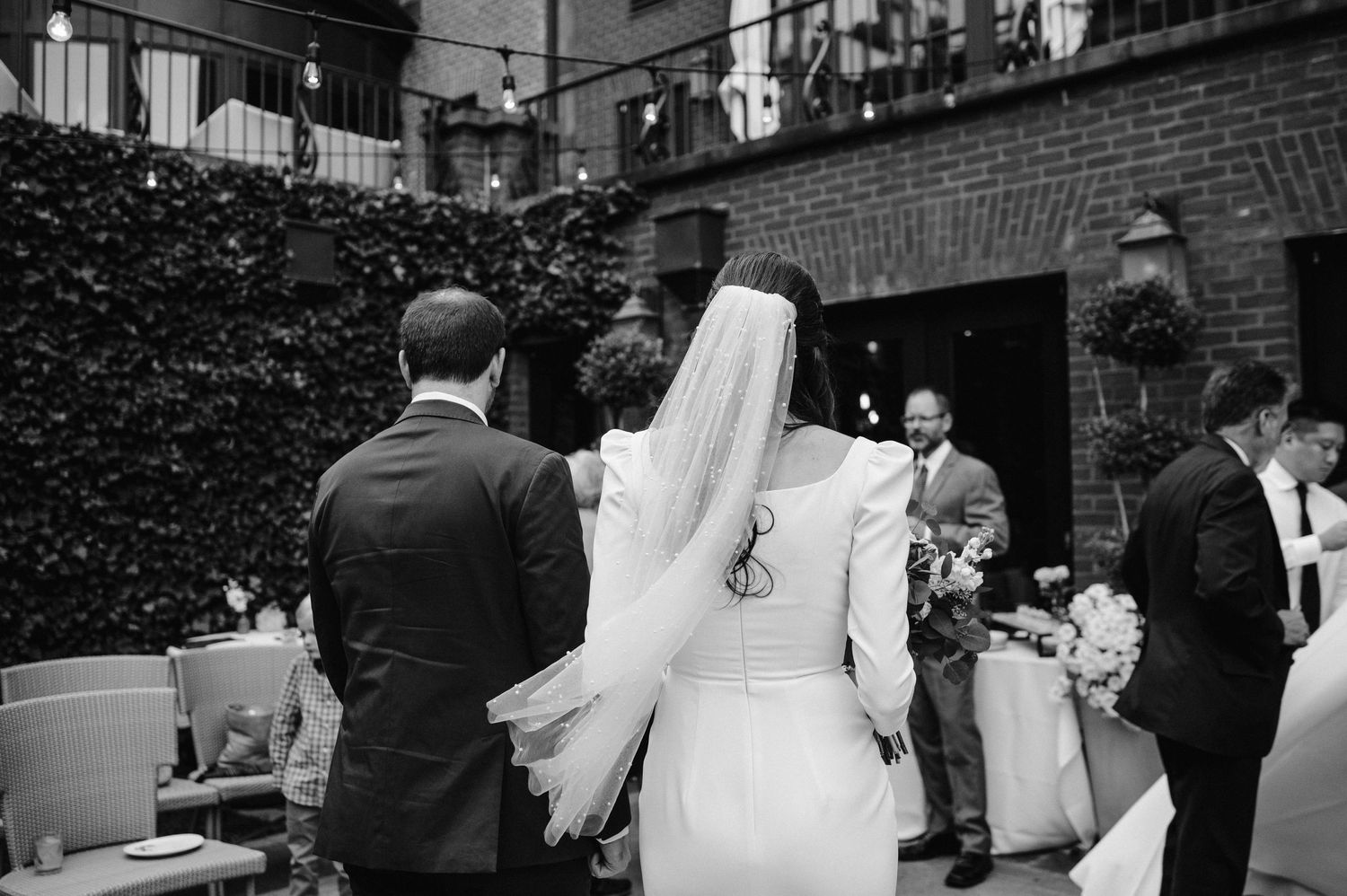 Black and white photo of a wedding reception with guests gathered in an ivy-covered courtyard.
