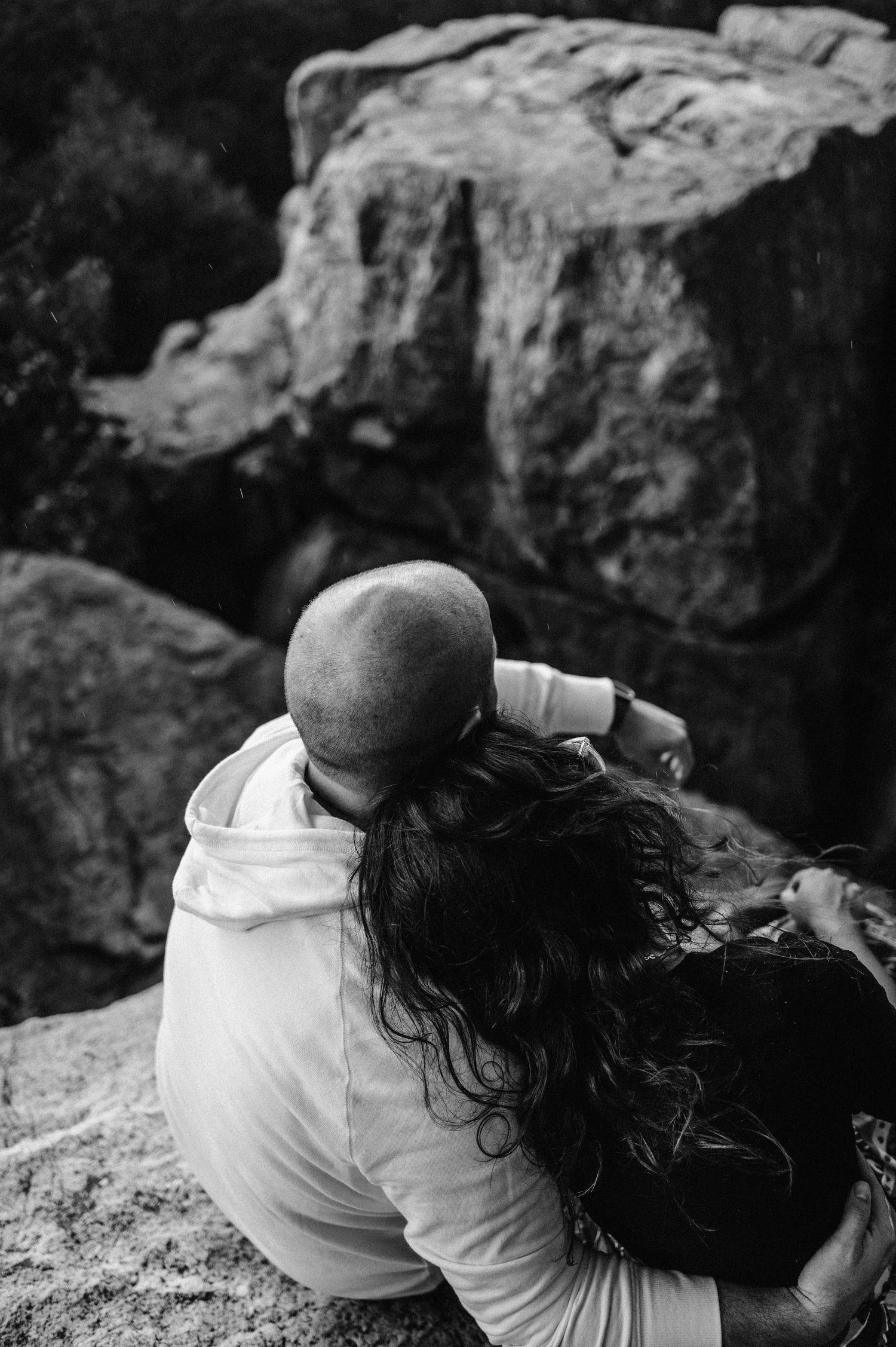 A black and white overhead view of a couple embracing while sitting on rocky mountain ledge.