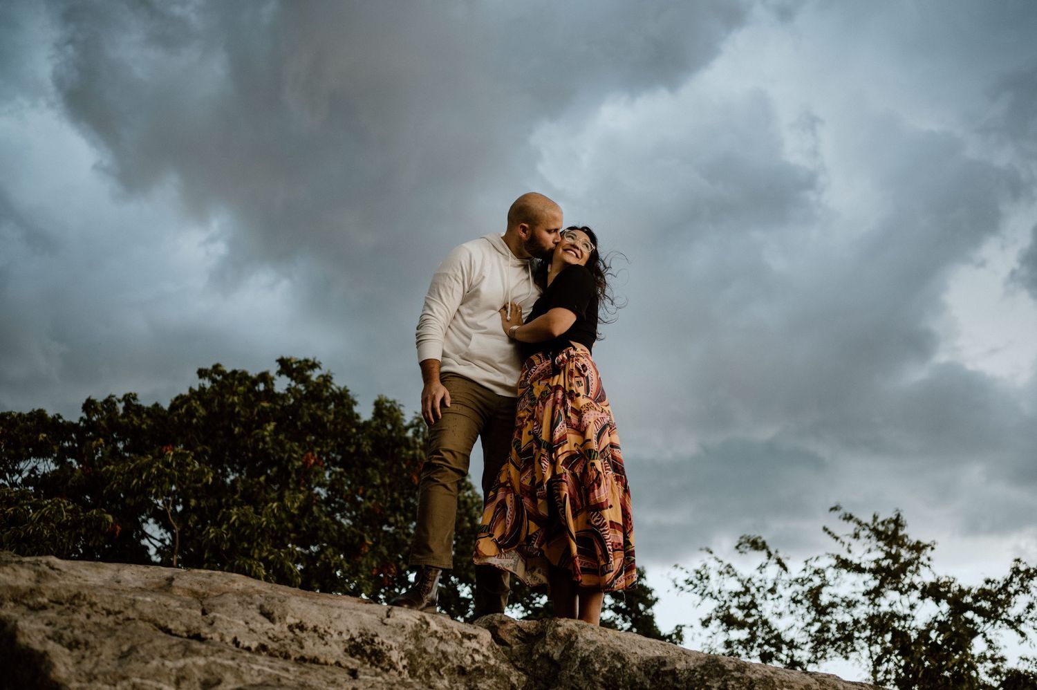 A couple stands together on rocky mountain terrain with moody storm clouds in the background.