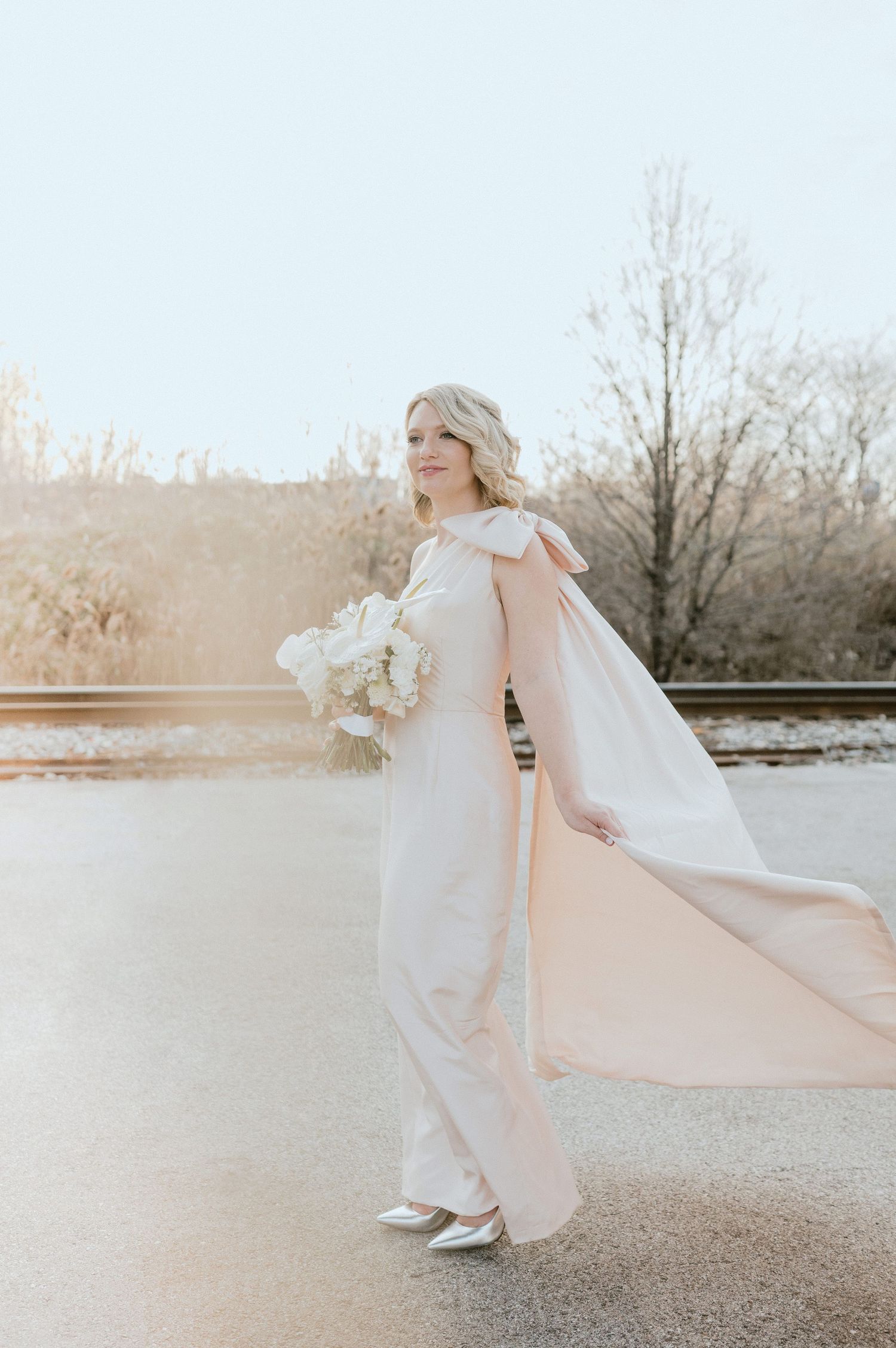 Bride in flowing white dress poses with bouquet against soft sunlit background.