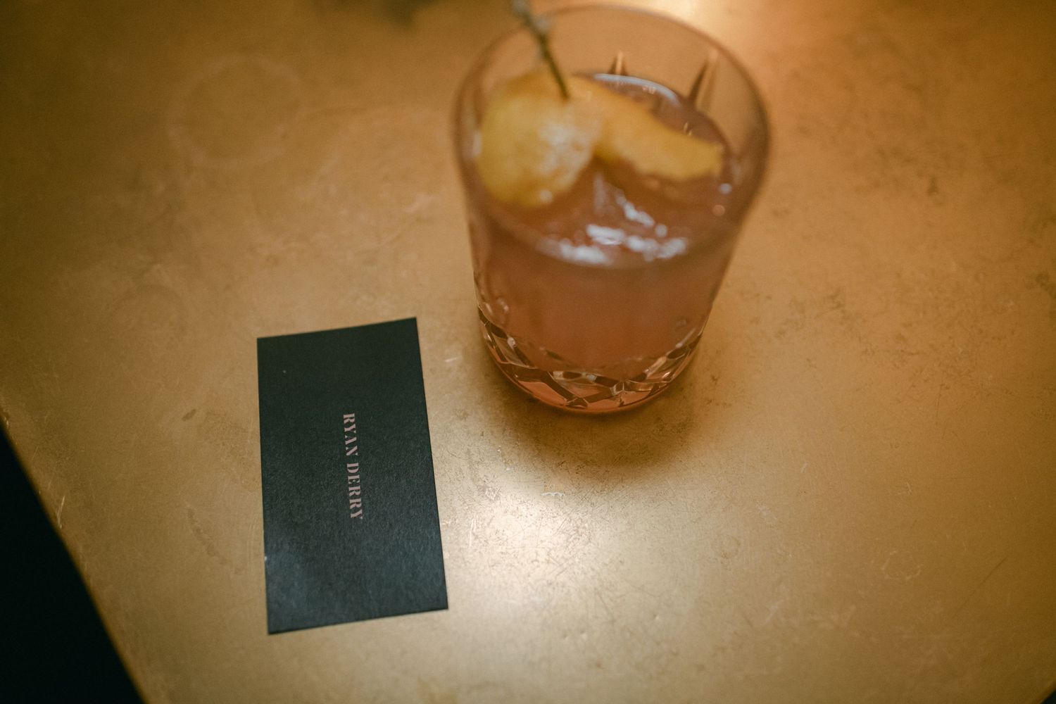 A cocktail glass and business card sitting on a wooden table surface.