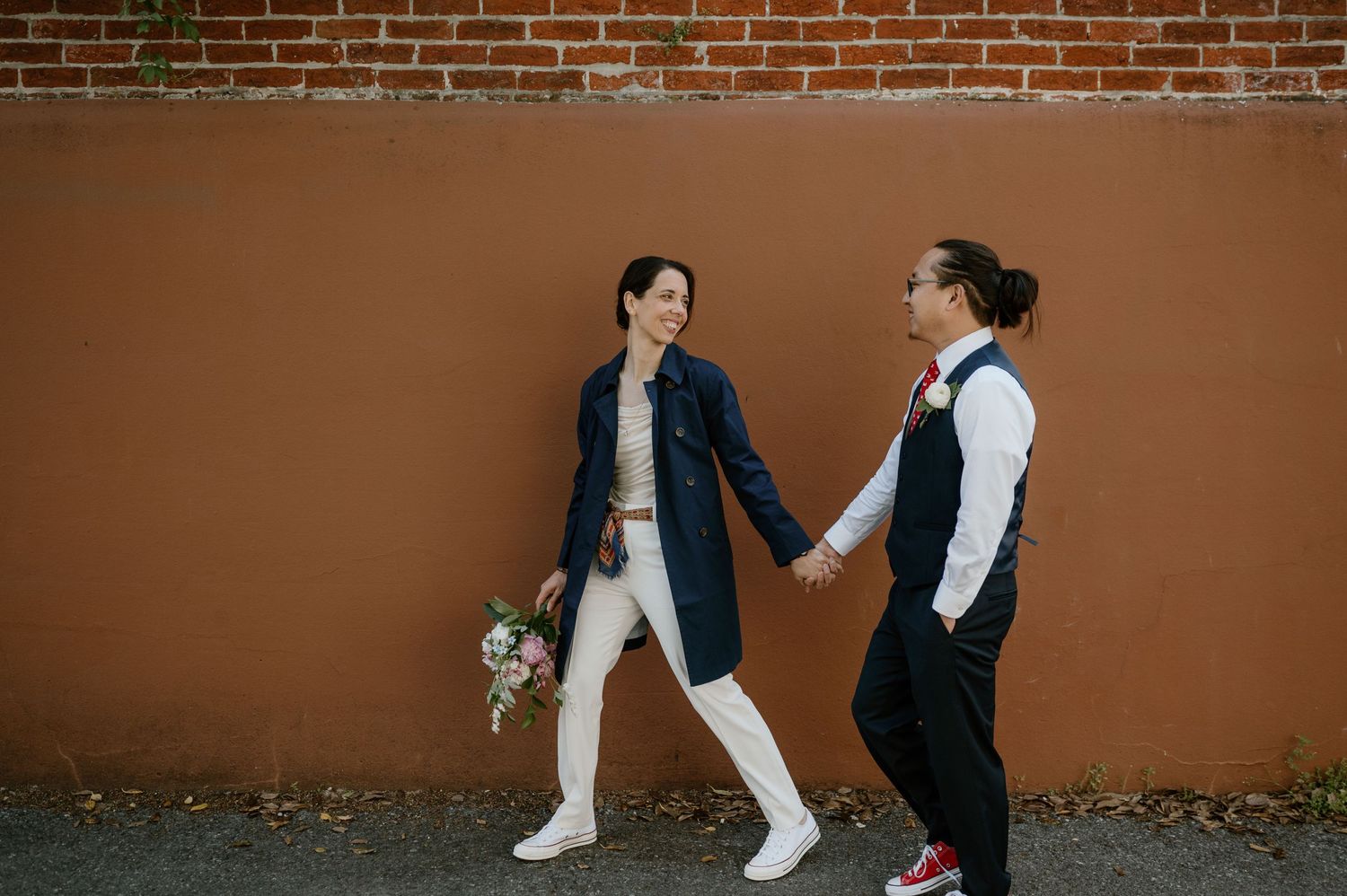 Two people in contrasting formal outfits walk together against a terracotta colored wall.