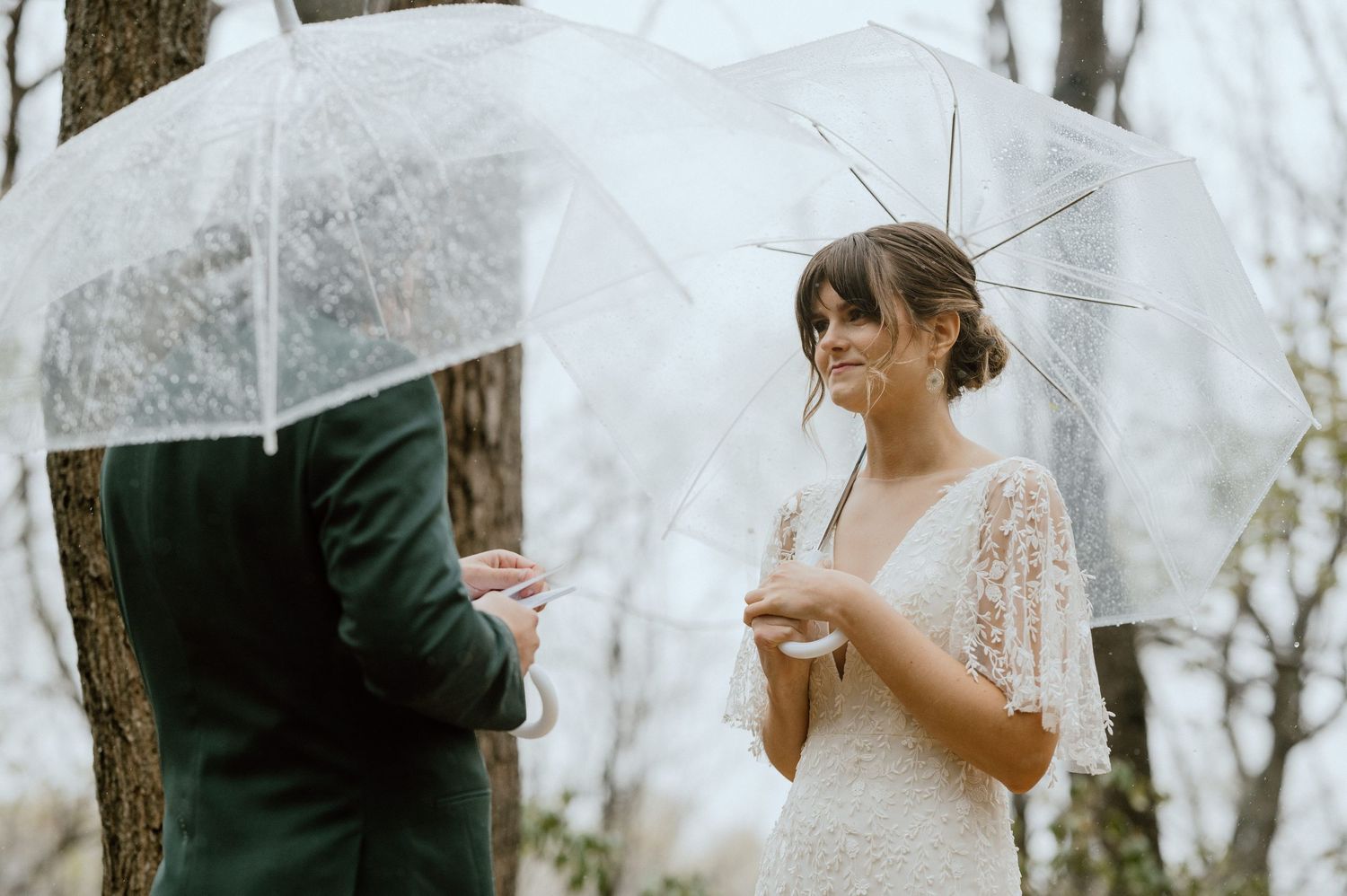 A couple shares an intimate moment under clear umbrellas in a wooded setting during their wedding ceremony.