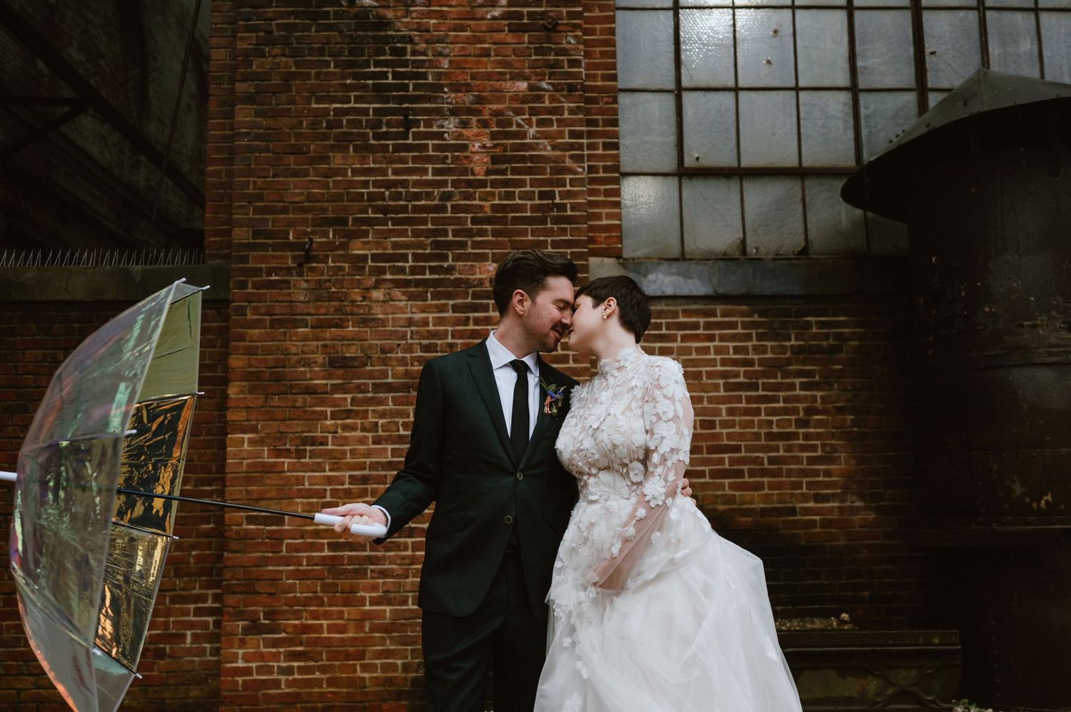 Couple shares romantic moment with transparent umbrella against industrial brick building.