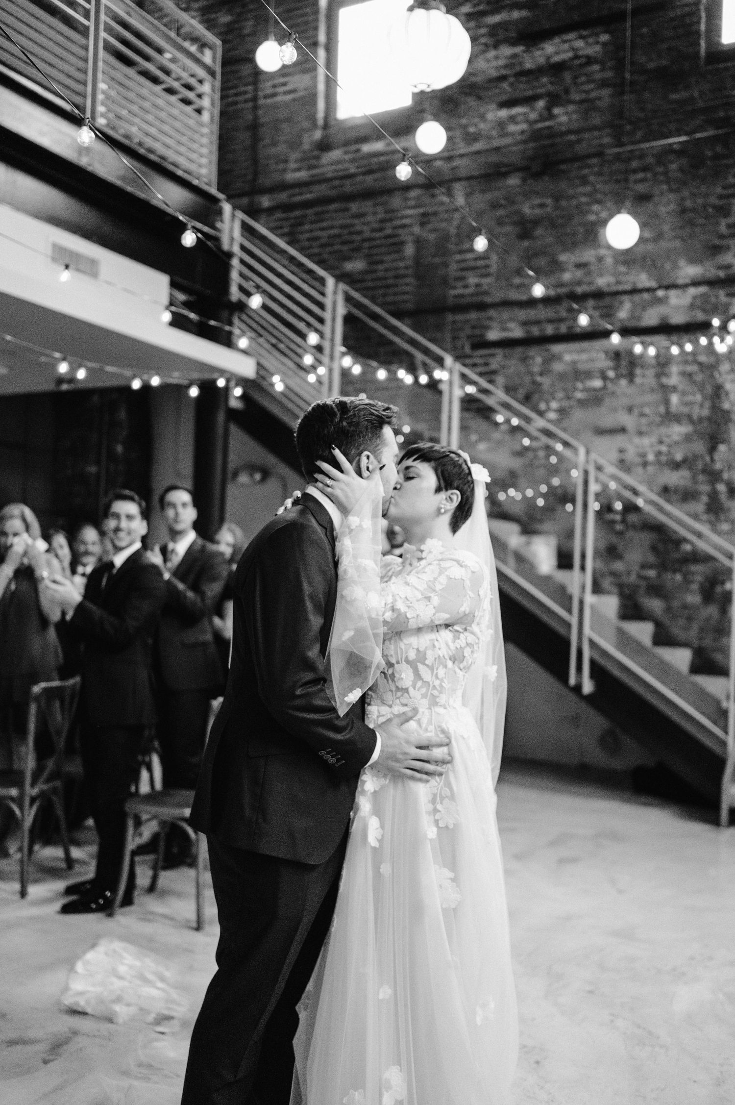 A newlywed couple shares an intimate kiss under string lights with exposed brick walls and staircase in background.