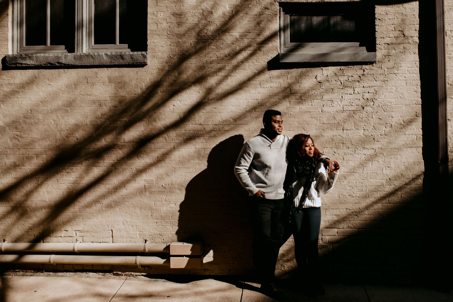 A couple stands against a brick wall with dramatic shadow patterns.