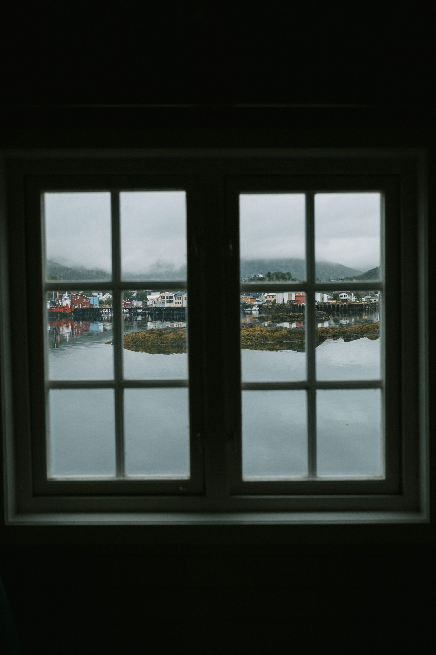 View through a window of a coastal village and mountains reflected in calm waters.