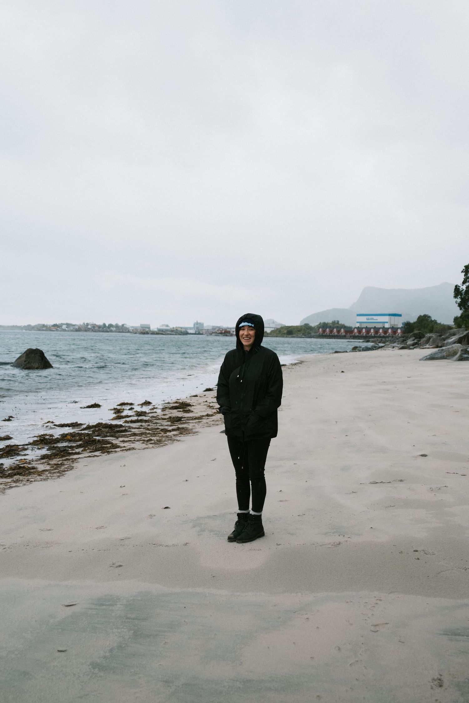 Figure in dark clothing stands alone on windy beach with cloudy coastal backdrop.