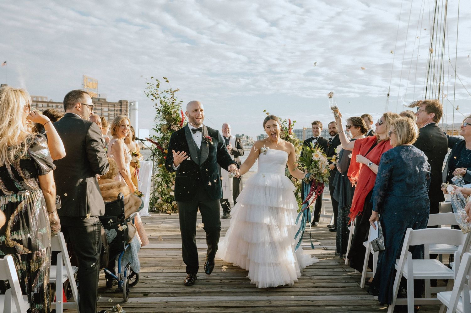 Newlyweds walk down a wooden dock pier as wedding guests celebrate and shower them with confetti.