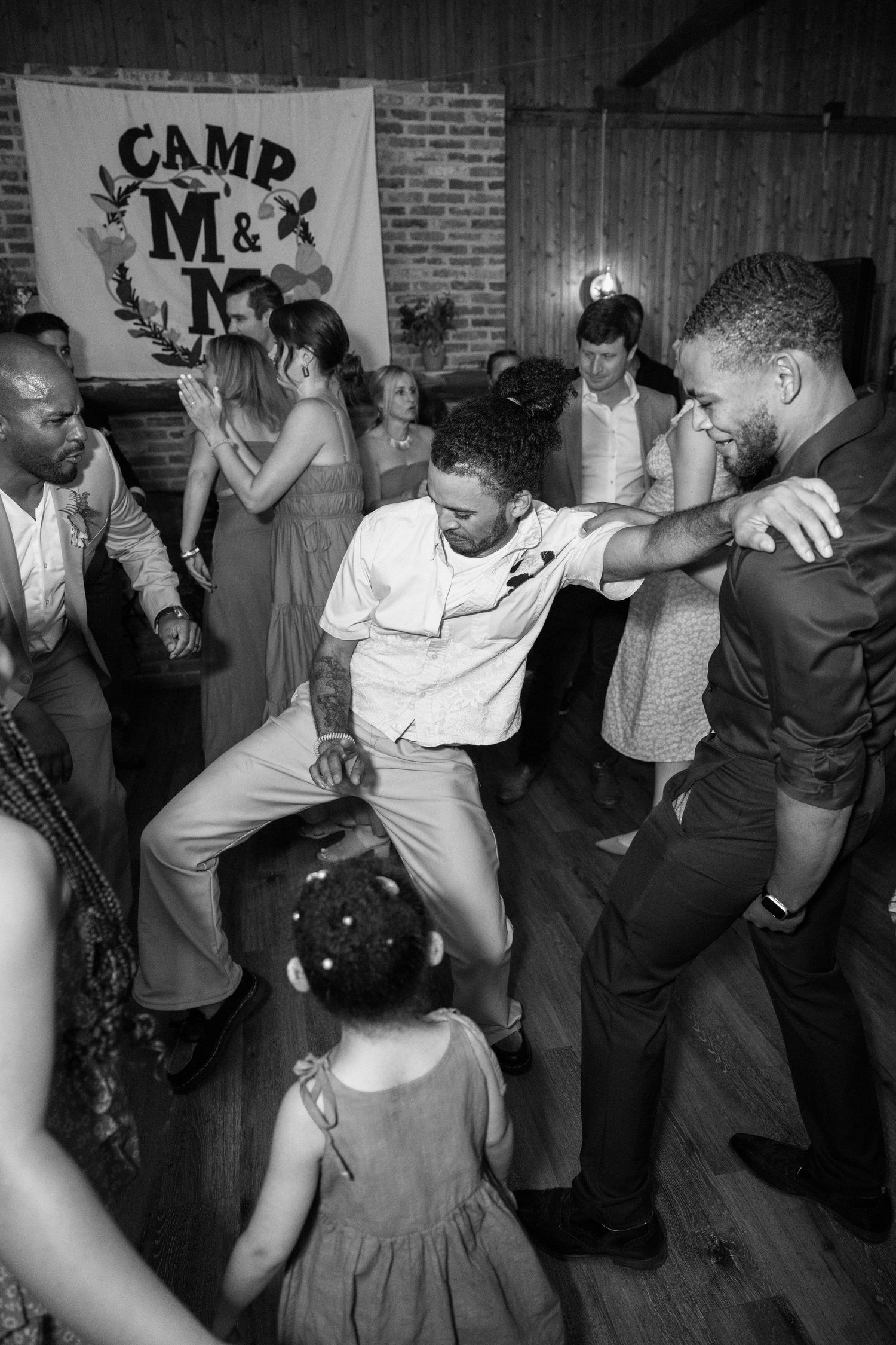 Energetic dance party at wedding reception with Camp M&M banner in background.
