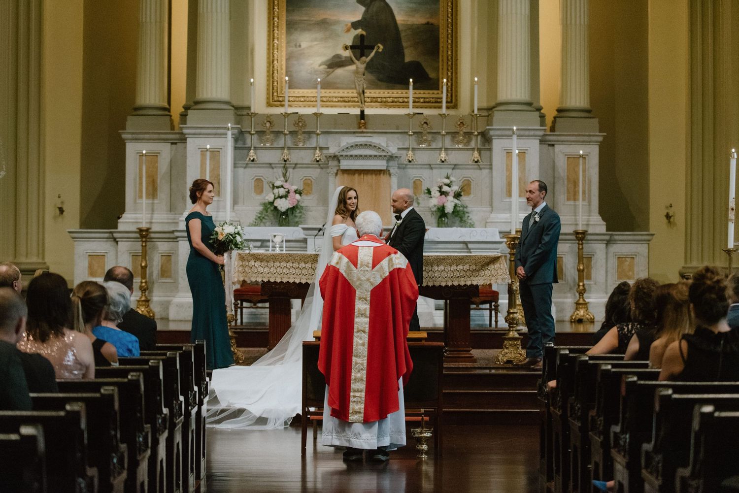 Catholic wedding ceremony with priest in red robes officiating at ornate church altar.