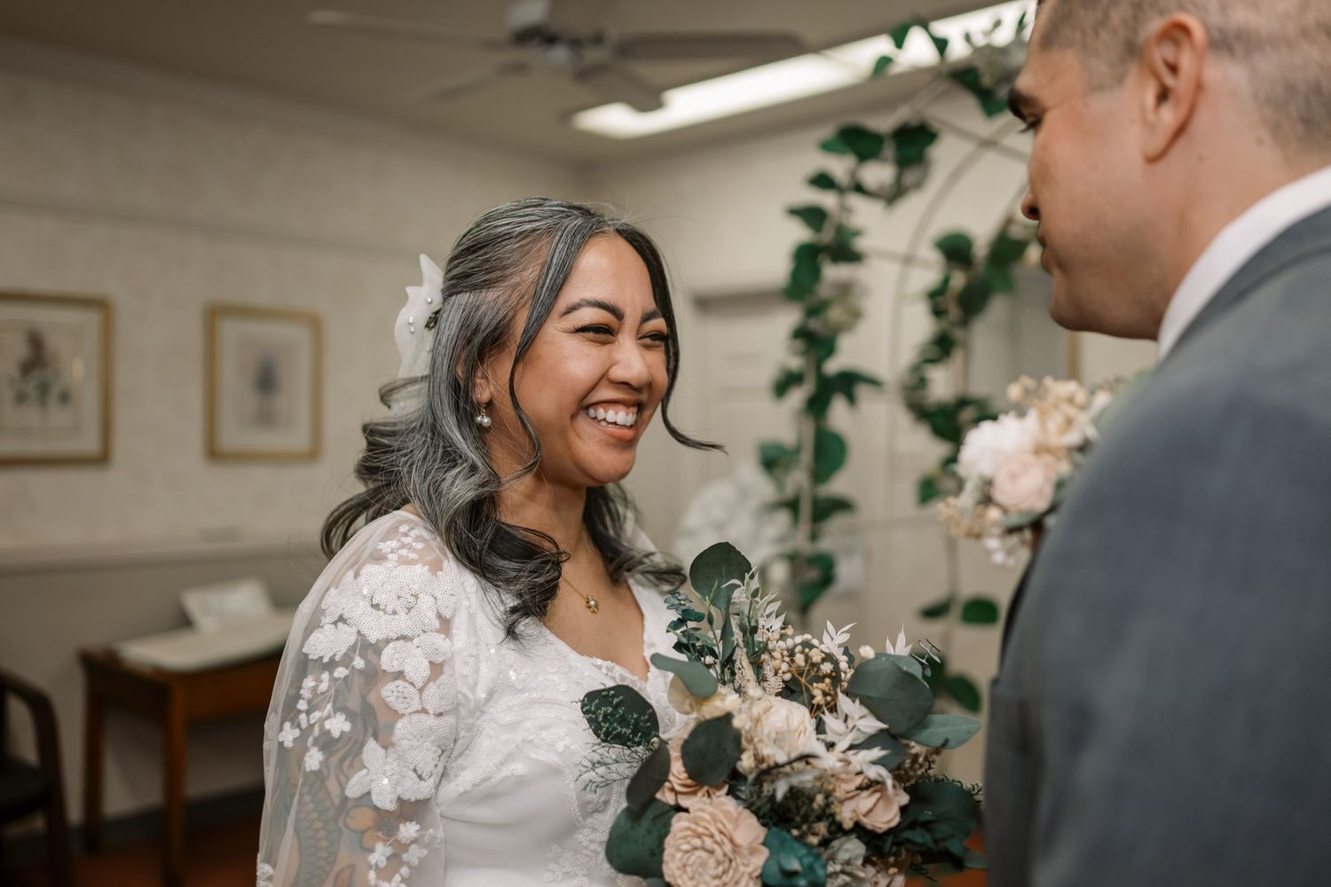 A bride in a white lace dress holds a bouquet of blush roses and eucalyptus during an indoor ceremony.