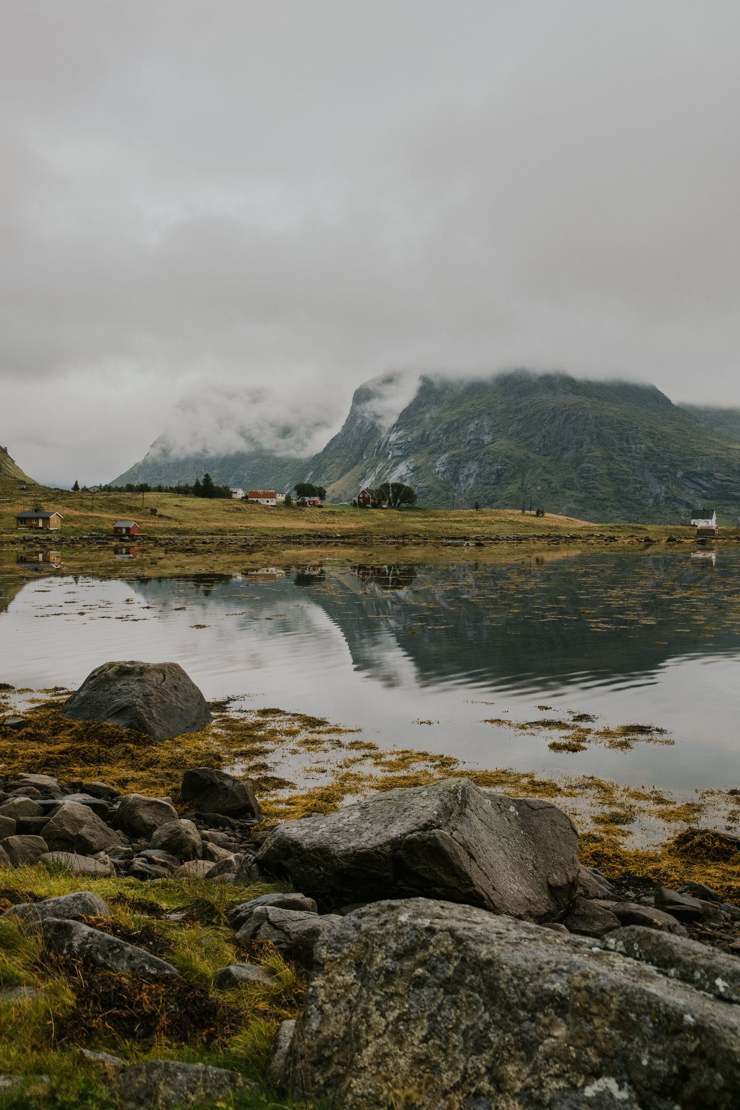 Rocky shoreline with mountain reflections in still fjord waters under cloudy sky.