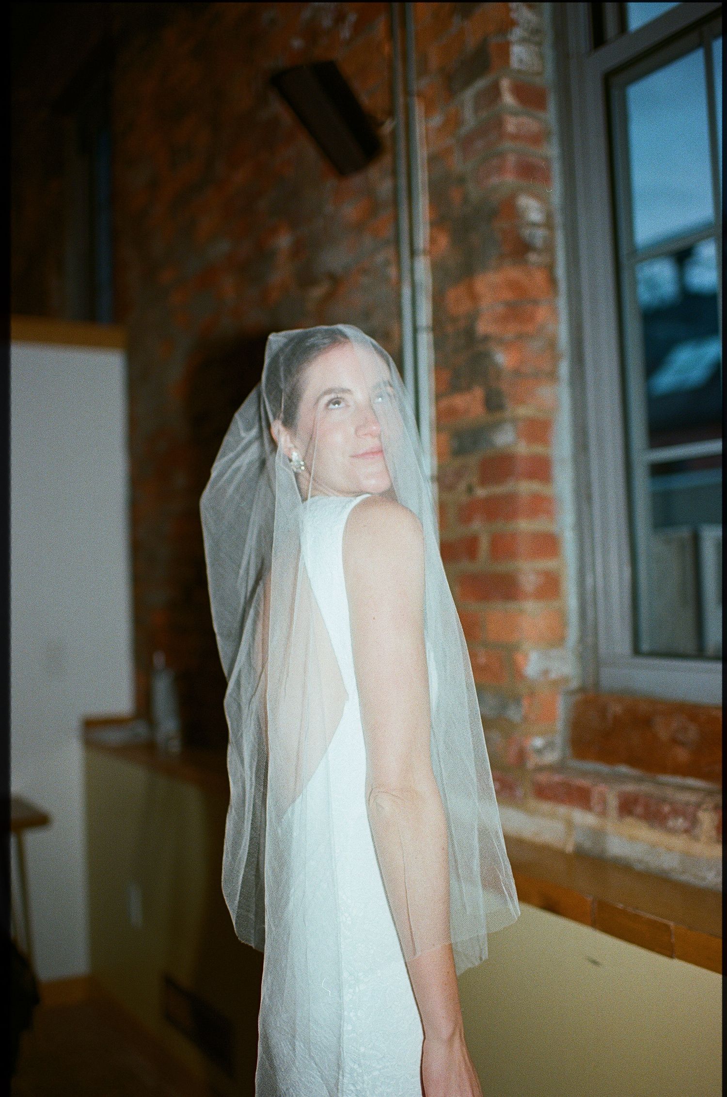 A bride in a white wedding gown and long veil stands by exposed brick walls in an indoor setting.