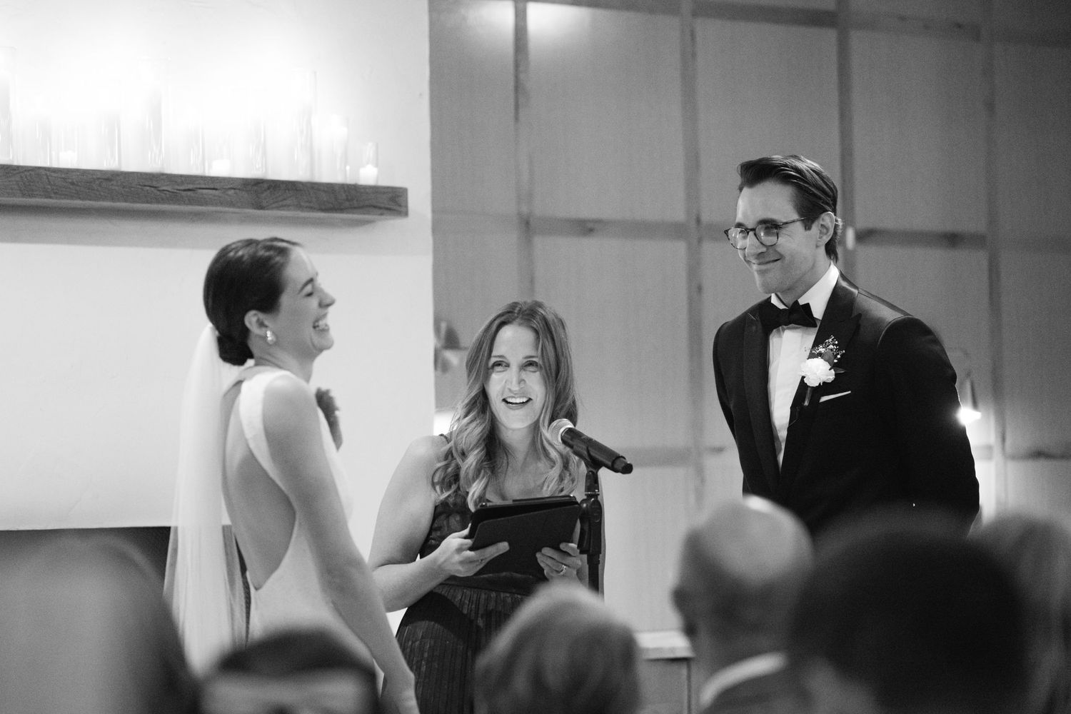 A wedding ceremony in progress with an officiant in black reading from a book as people gather in black and white.