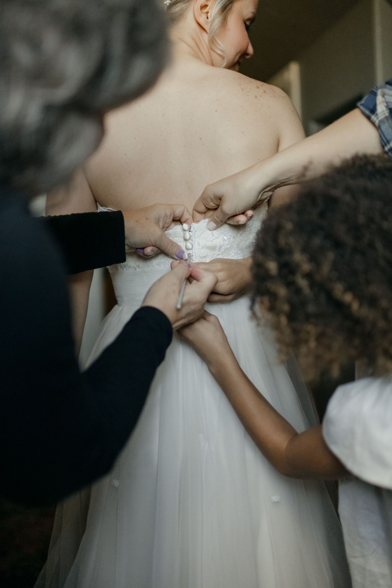 Multiple hands working together to fasten buttons on back of white wedding gown.