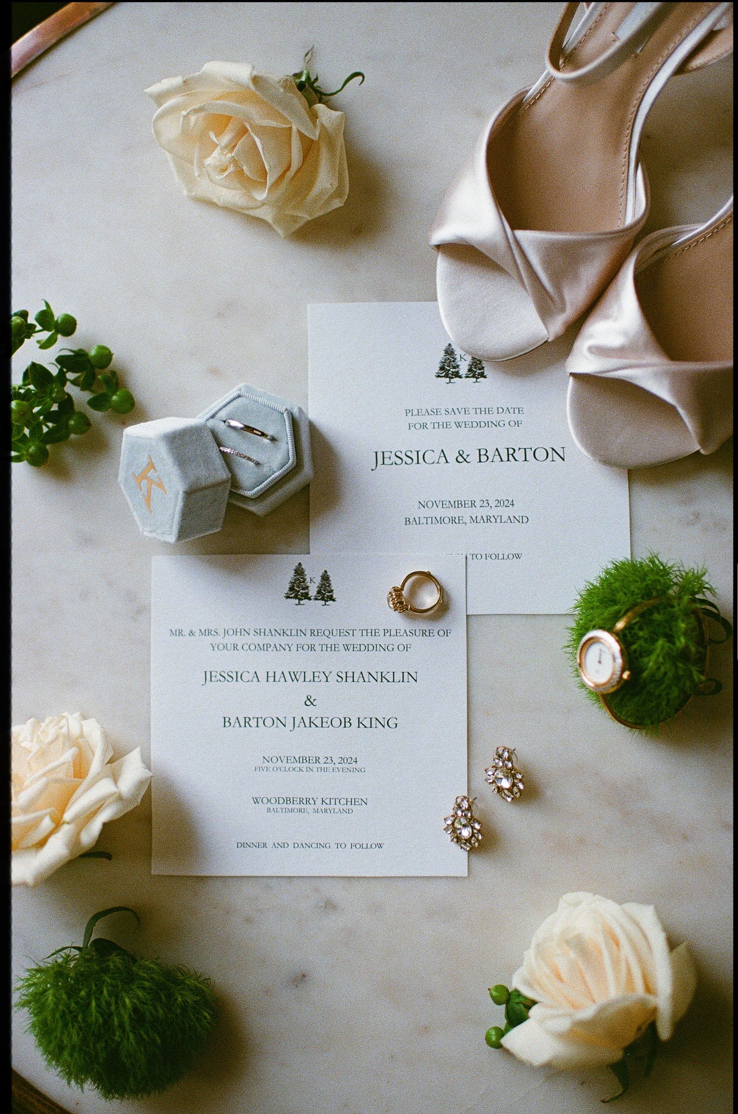 Elegant wedding invitation with white roses, green accents, and bridal shoes on marble surface.
