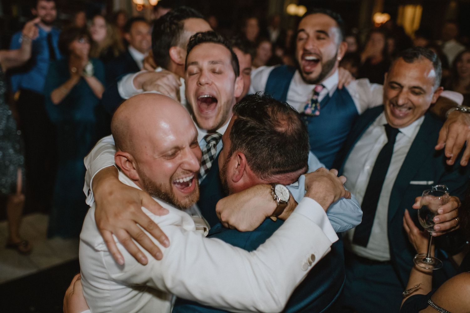Group of wedding guests embrace and celebrate with joyful expressions at indoor reception.