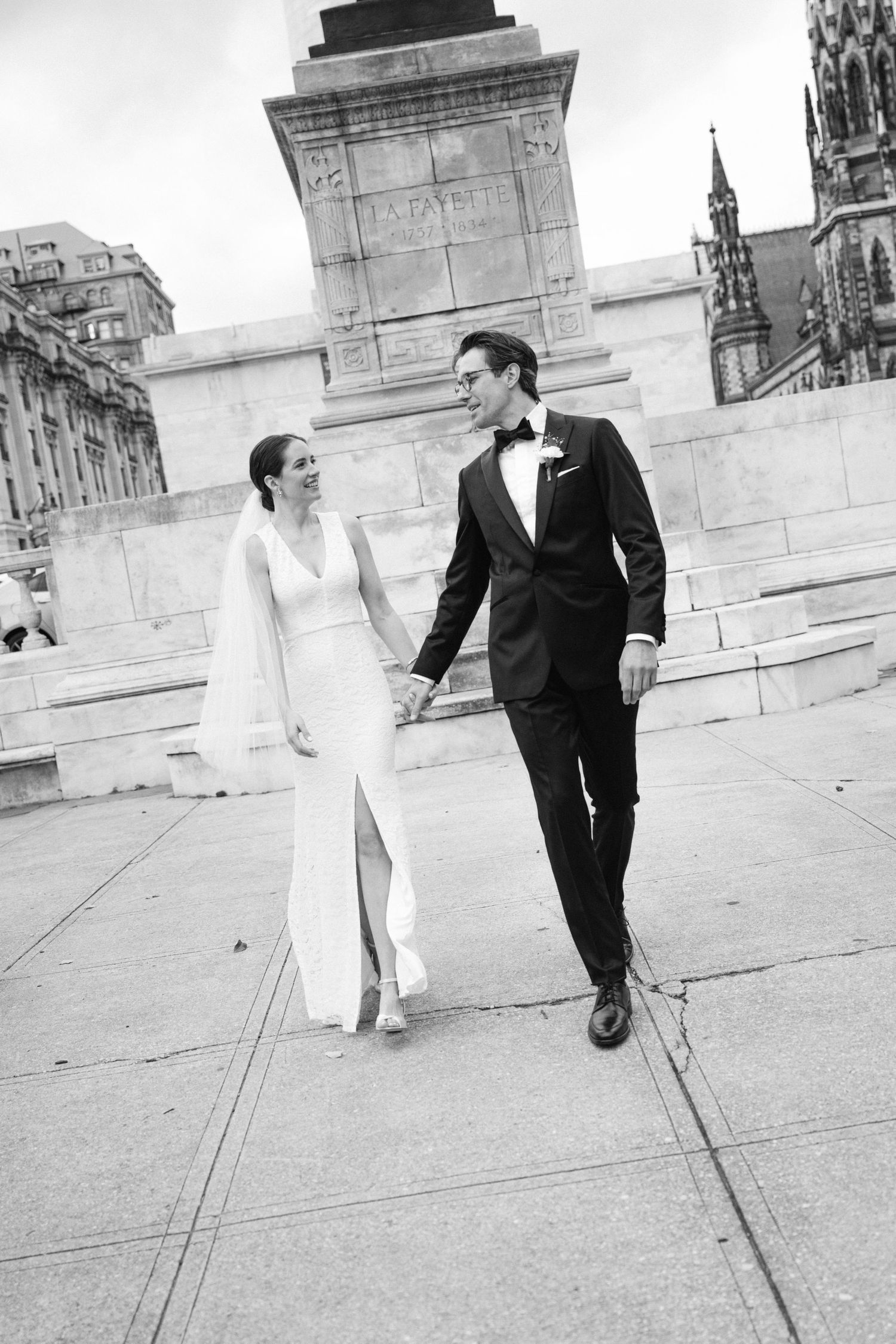 Elegant couple in formal attire walking along stone architecture in black and white.