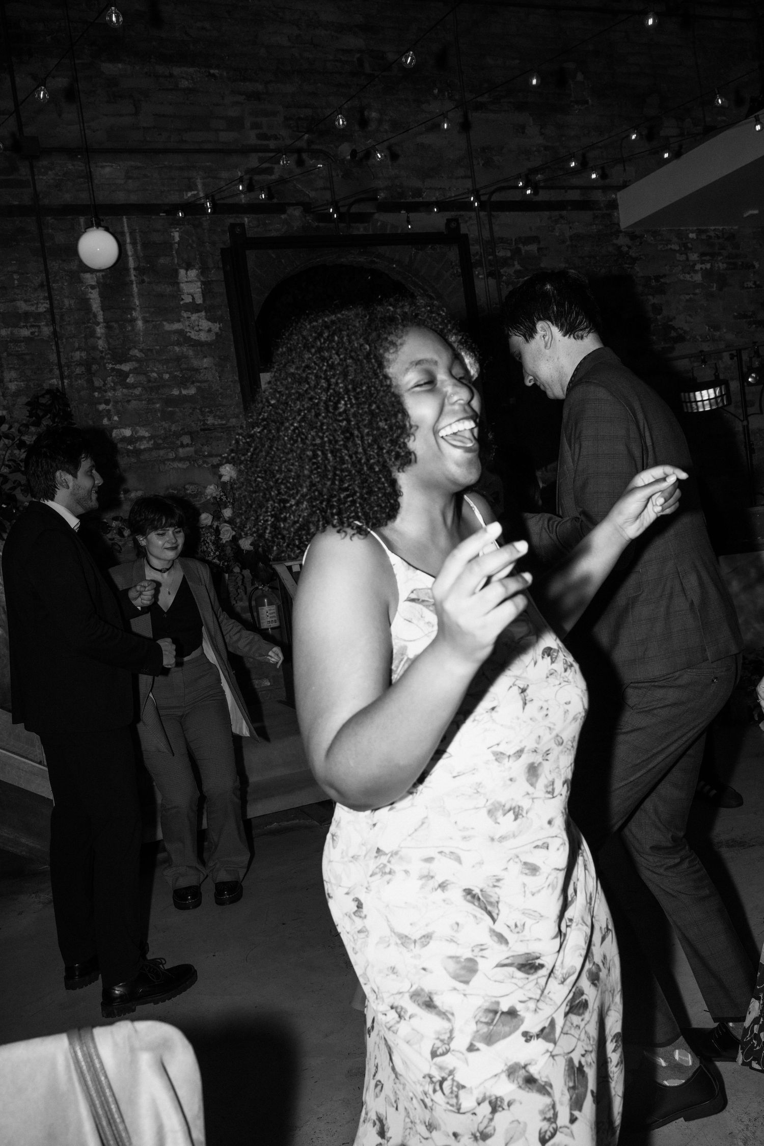 Woman in floral dress dancing joyfully at a party with exposed brick walls and dim lighting.
