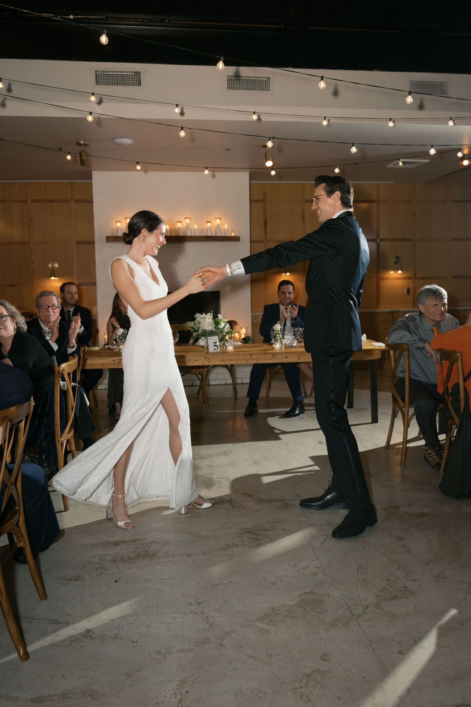 A couple shares their first dance in an intimate venue with string lights and wooden decor.