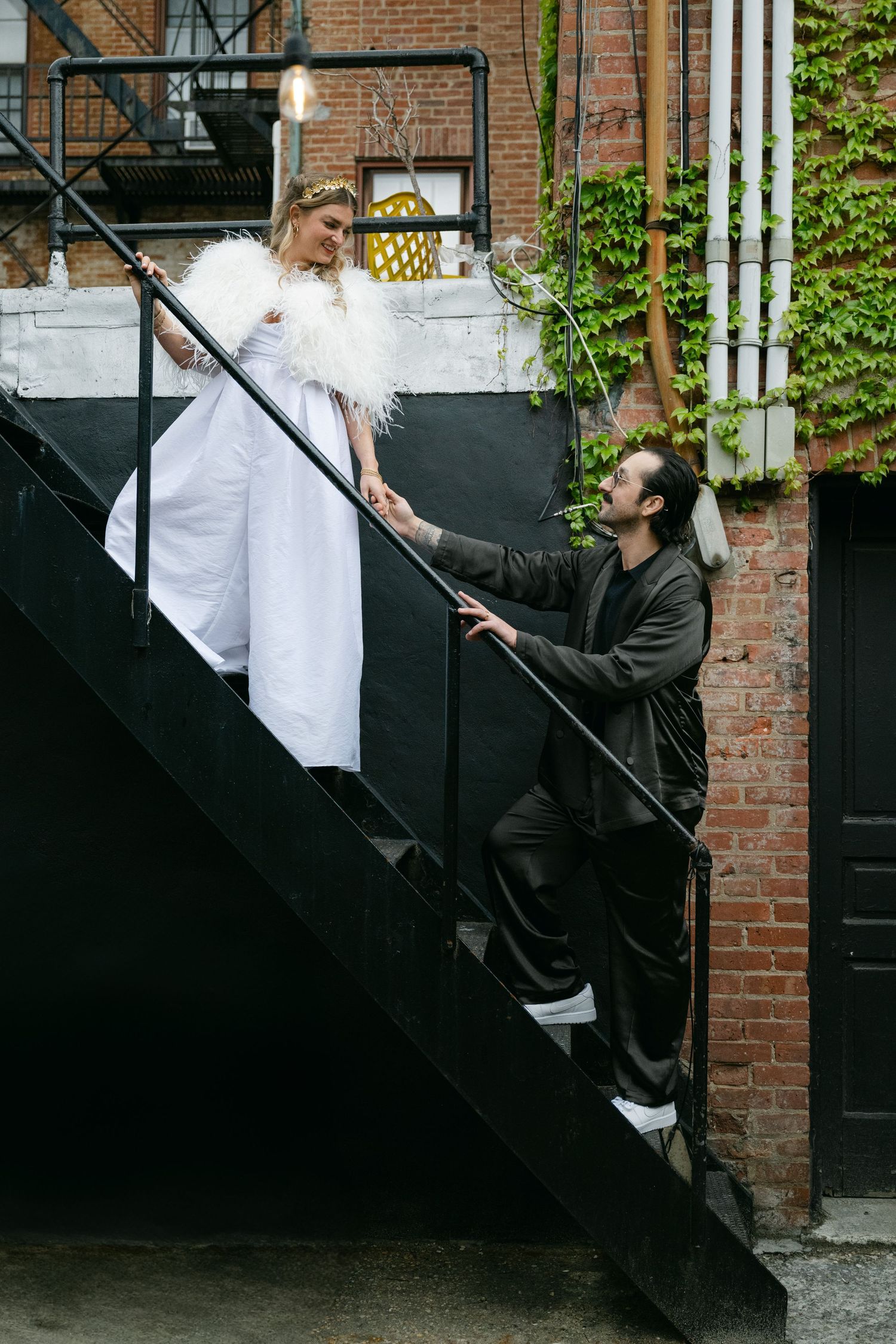 A couple poses playfully on black metal fire escape stairs against a brick building exterior with string lights at Fadensonnen in Baltimore