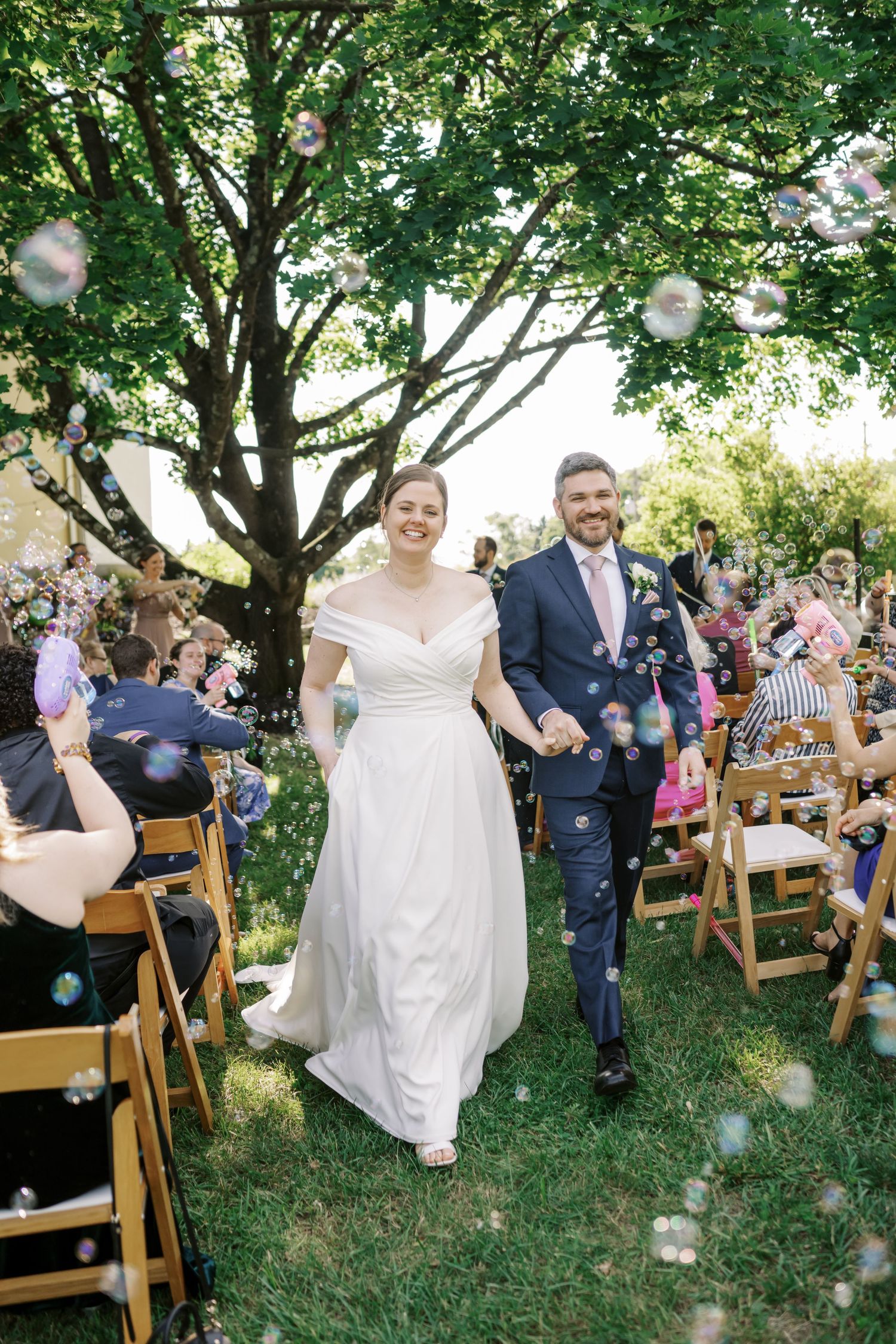 A joyful outdoor wedding ceremony with bubbles floating through the air under a large tree.