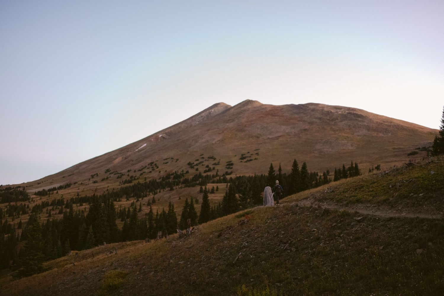 Rocky mountain peak rises above evergreen forest during sunset in a serene wilderness landscape.
