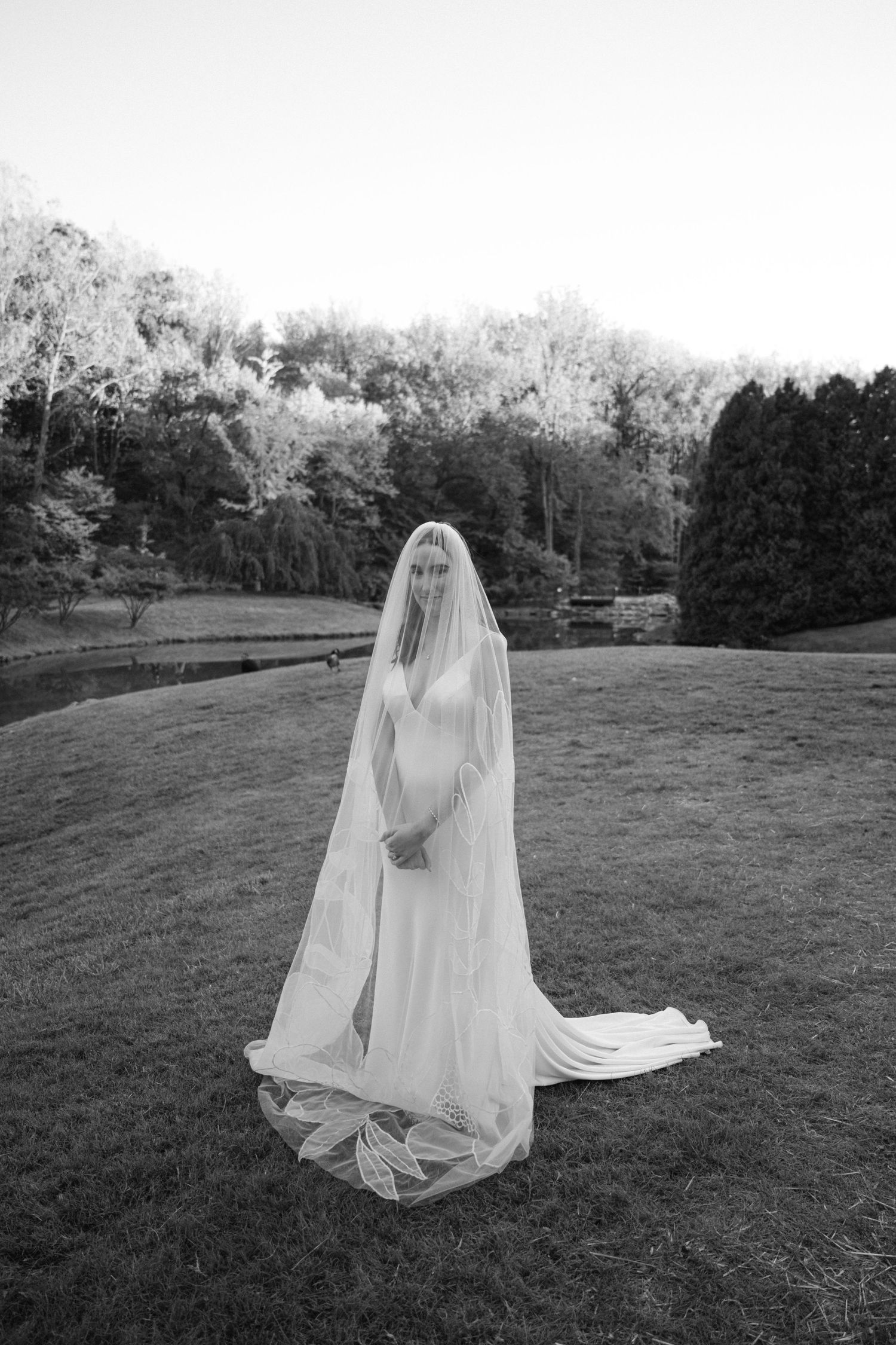 Black and white photo of a bride in a flowing wedding dress and veil standing in a grassy field.