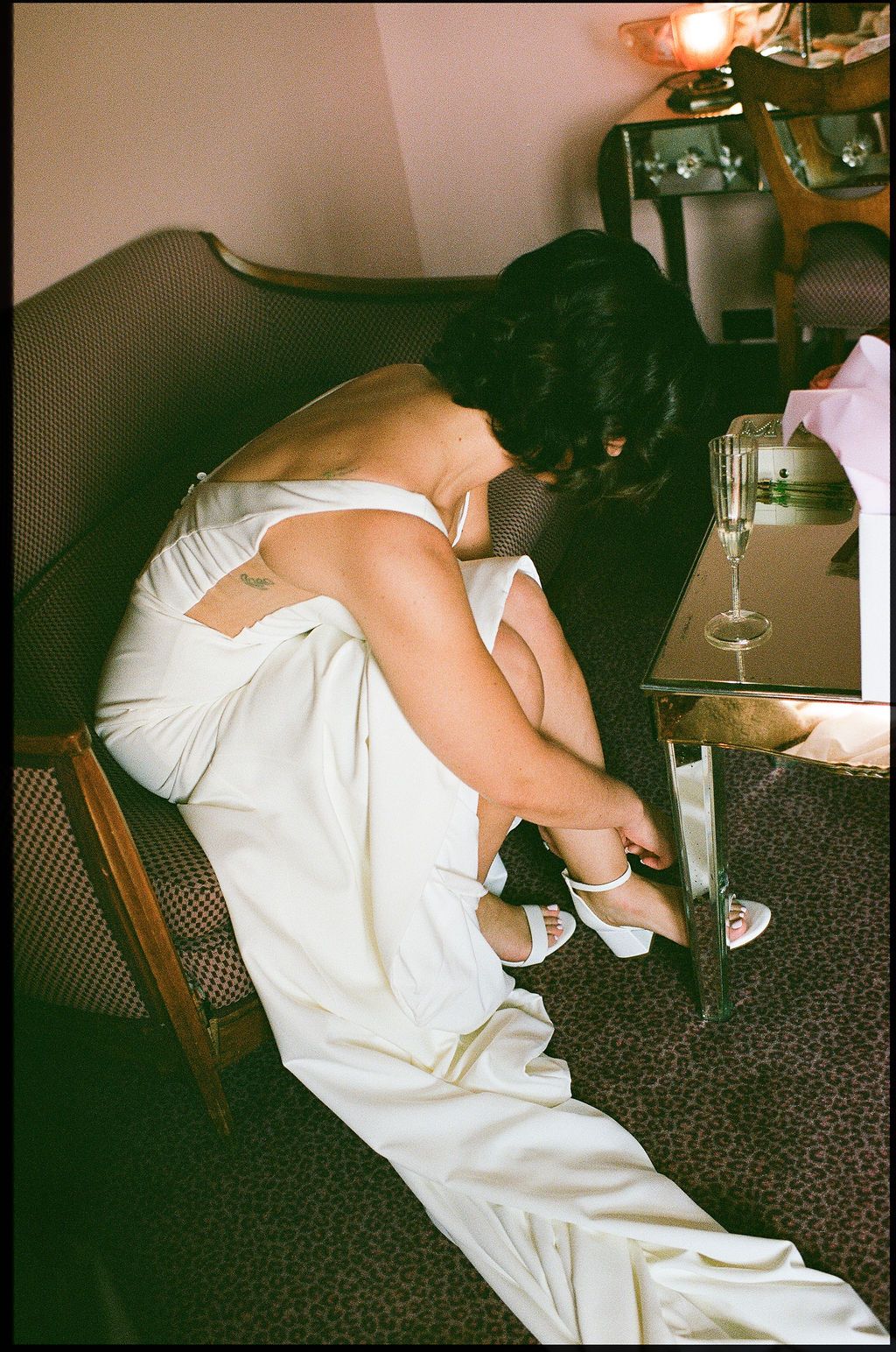 A person in a white dress adjusts their white heels while seated on a dark couch in a dimly lit room.