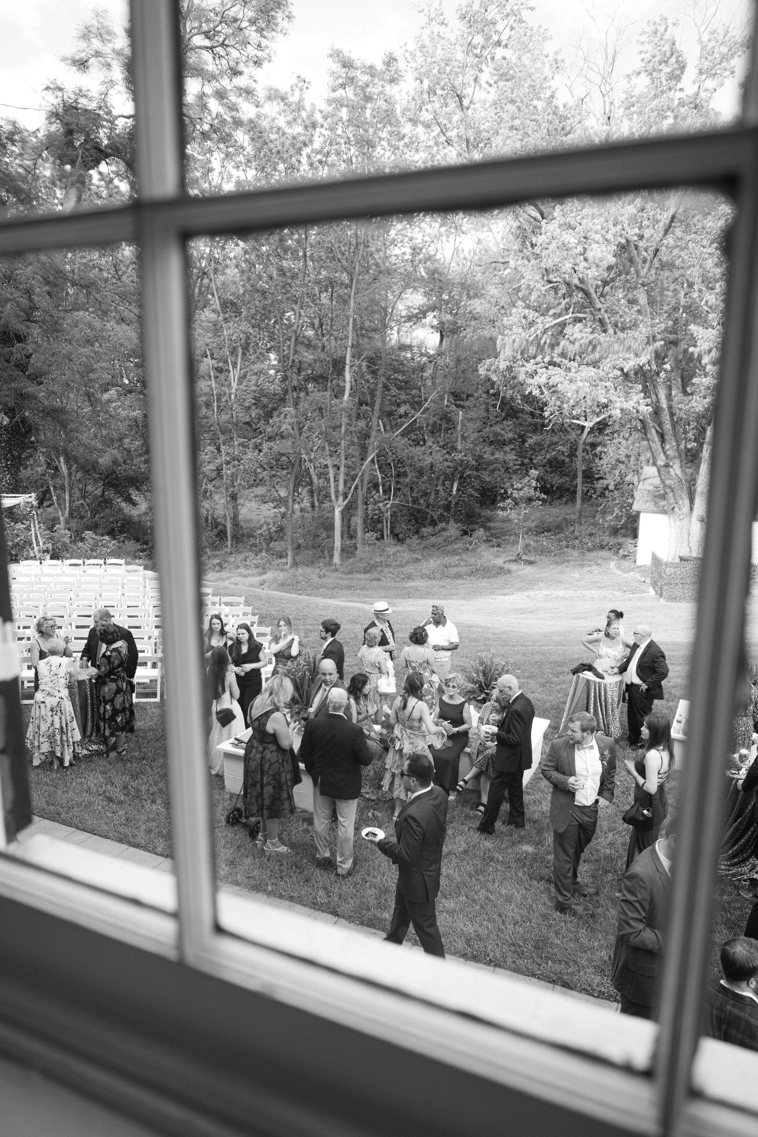 A line of wedding guests gather outdoors on a grassy slope viewed through window panes.