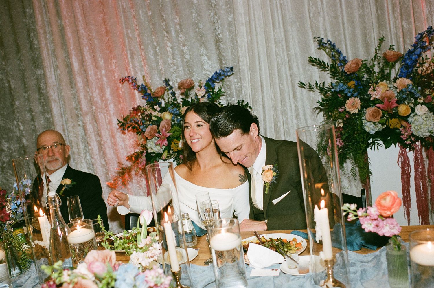 Bride and groom seated at floral-adorned wedding reception table with candles and colorful blooms.