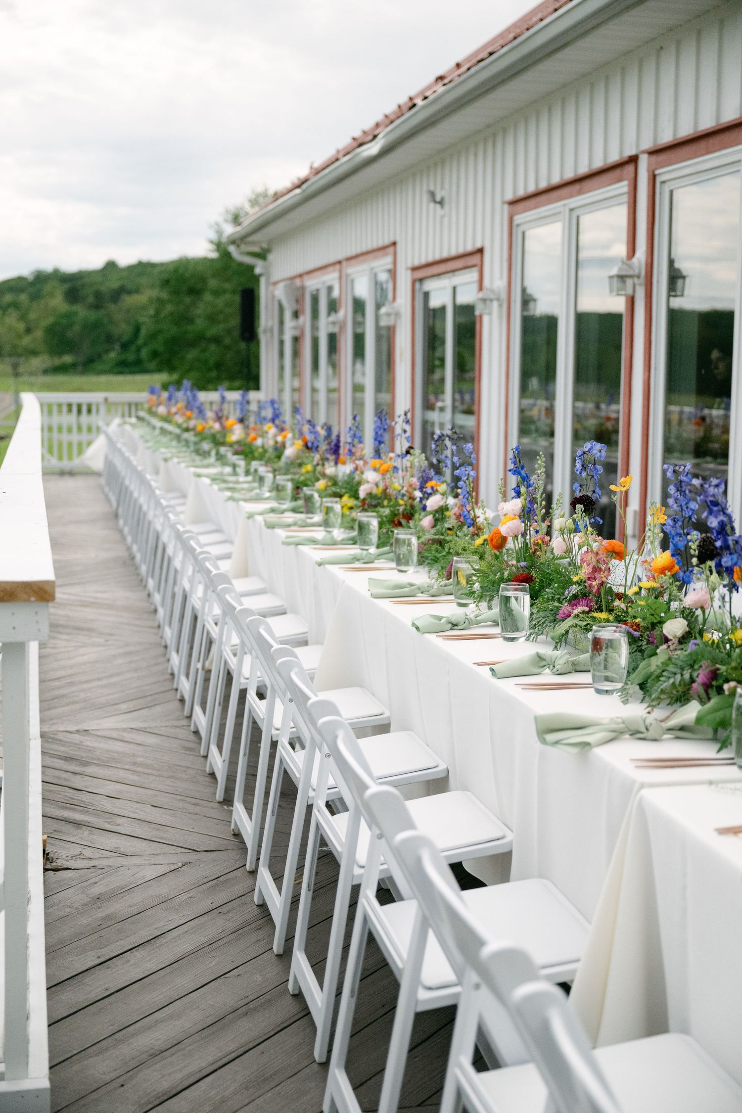 Long white reception tables with floral centerpieces set up on an outdoor deck.