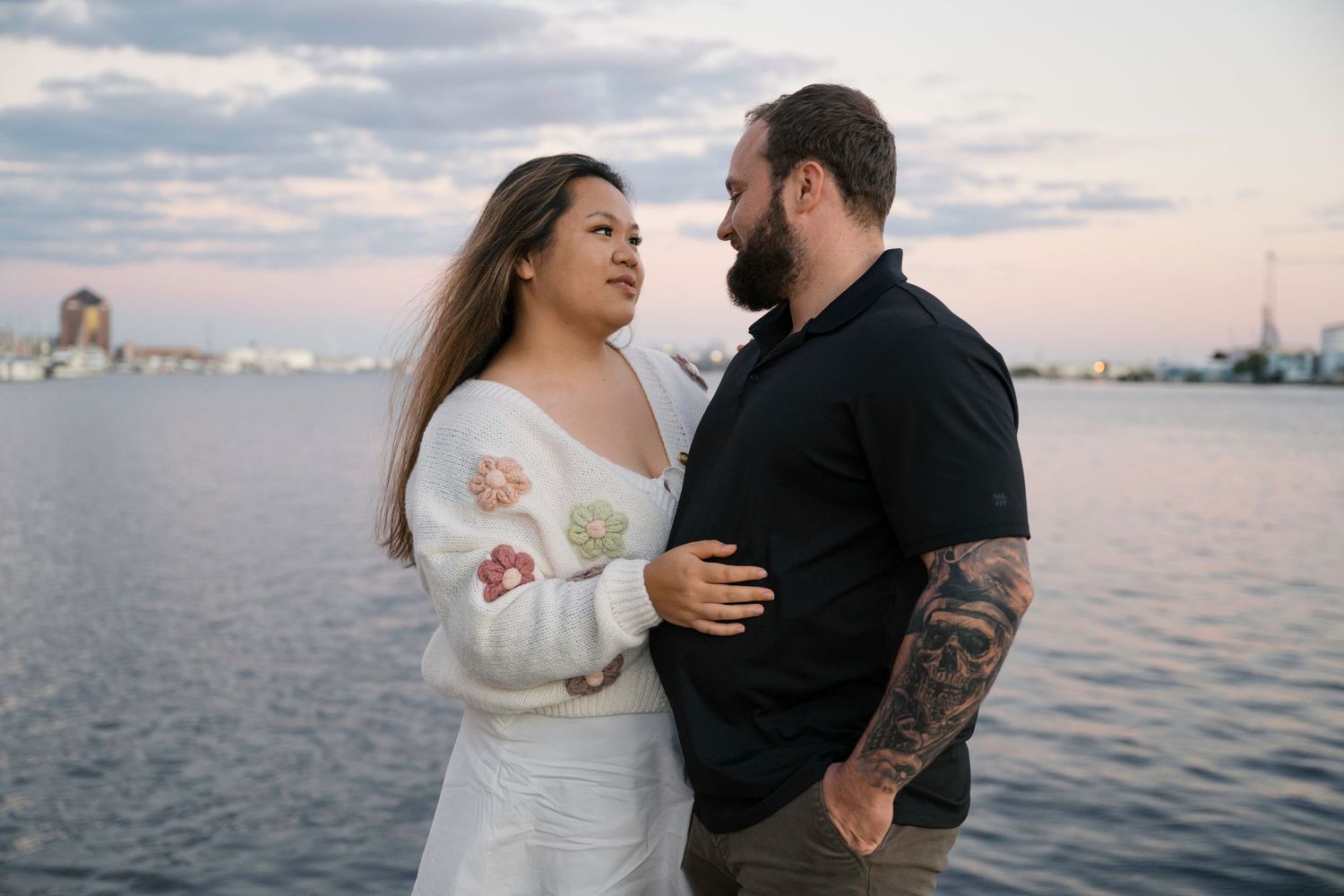 A couple embraces lovingly by the waterfront at sunset while boats dot the harbor in the distance.