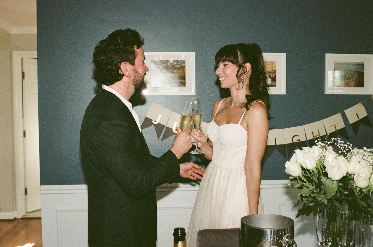 A couple raises champagne glasses in celebration, with a banner and white flowers decorating the teal-colored wall behind them.