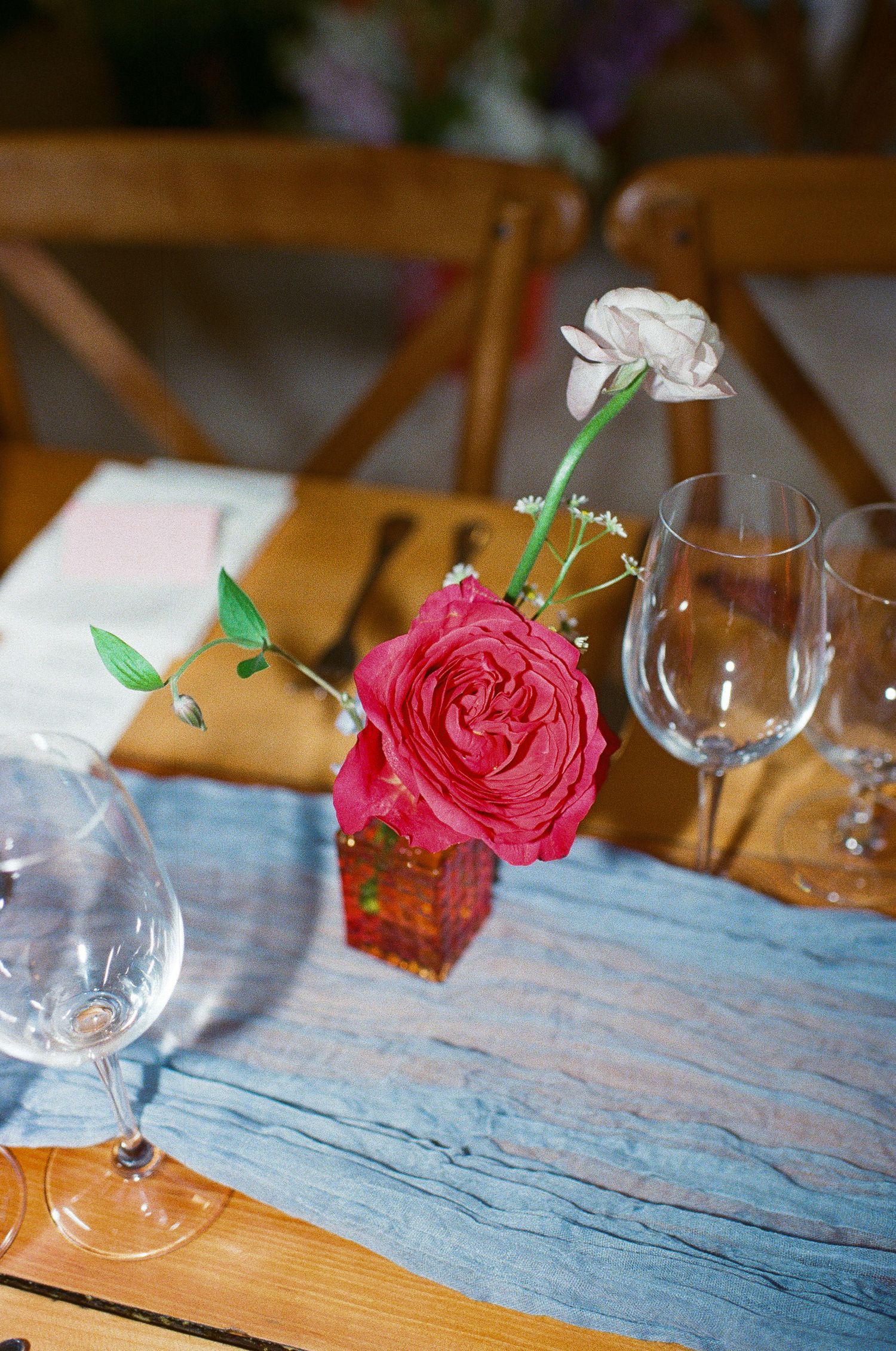 A single pink rose centerpiece in a small vase with wine glasses on a blue table runner.