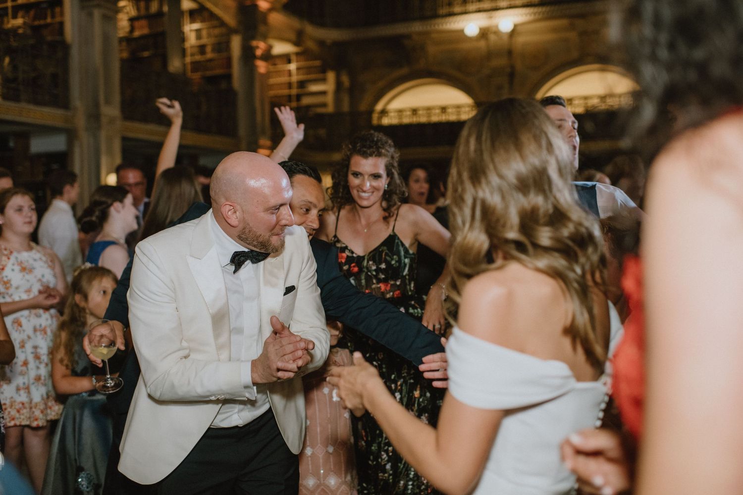 Wedding guests dance and celebrate in an elegant venue with dramatic lighting and architectural details.