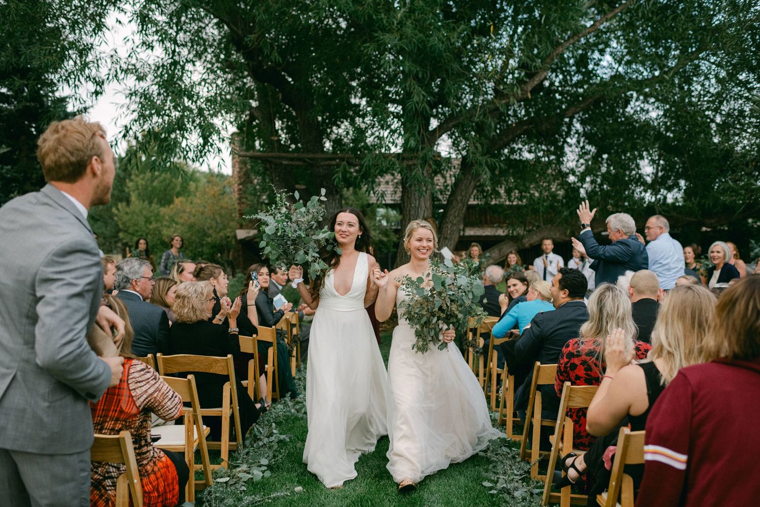 Wedding ceremony guests watch as two people in white dresses walk down an outdoor aisle together.