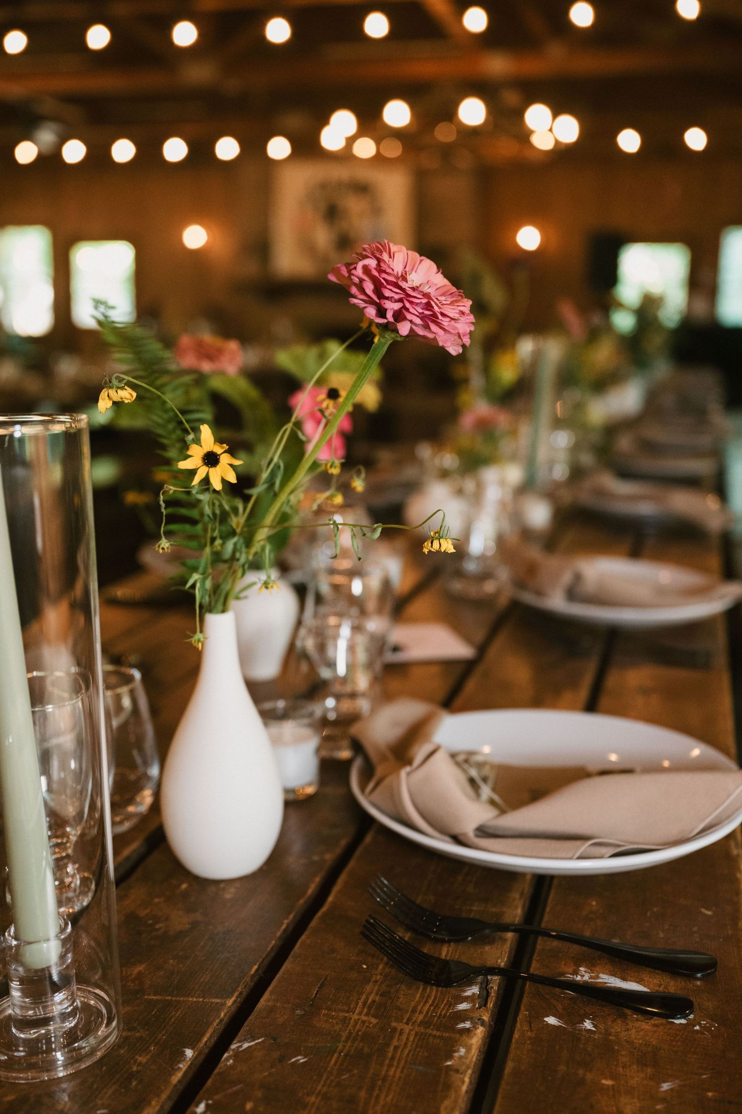 Rustic table setting with white vase holding pink flowers and glassware under warm string lights.